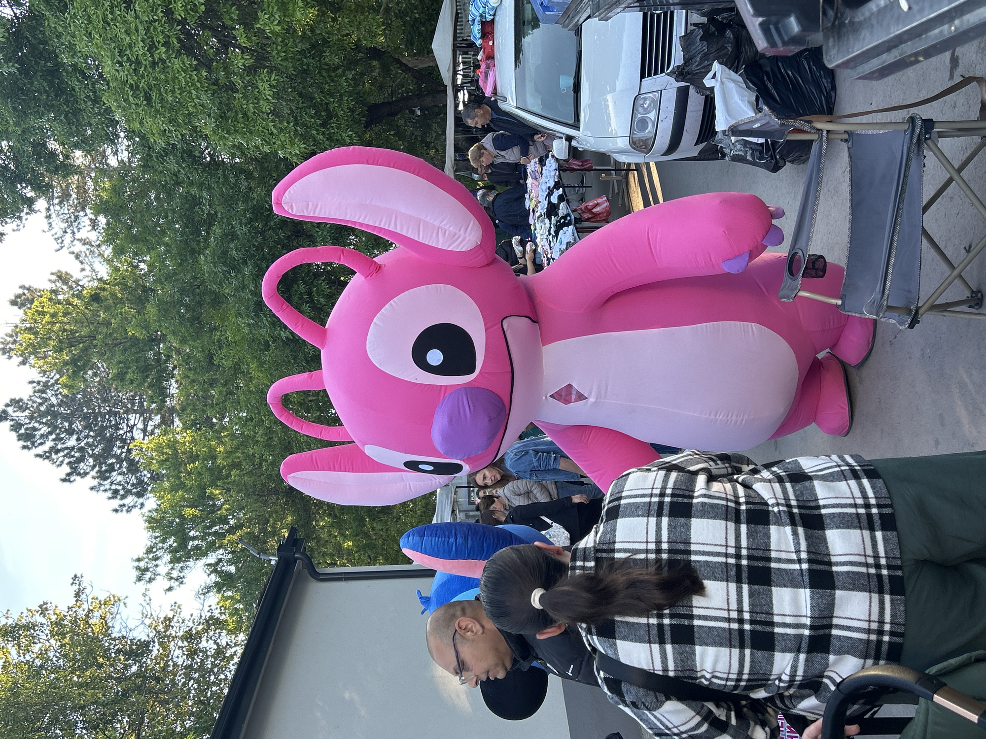 The image shows a pink inflatable figure resembling the Disney character Angel, positioned on what appears to be a sidewalk or paved area. Several people are standing nearby, including individuals wearing casual clothing such as a black and white plaid jacket and a black jacket. A white car is visible in the background, along with what seems to be a display table with various items on it. Green trees are also present, suggesting the scene is outdoors, possibly at a market or event. Based on the prompt, the location is Targovishte, Bulgaria.