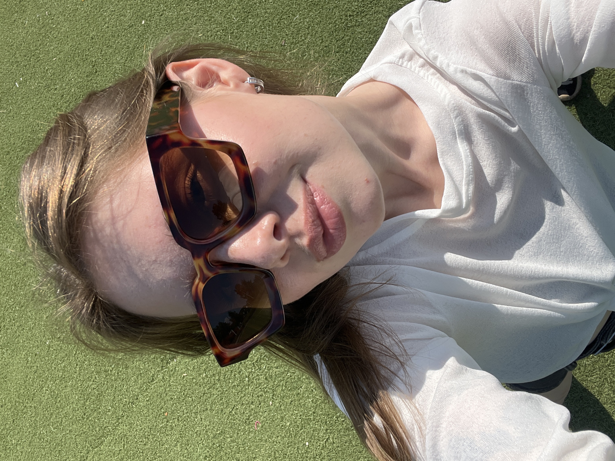 A young woman is taking a selfie outdoors on a sunny day. She is wearing large, tortoise-shell patterned sunglasses and a white long-sleeved shirt. She has light brown hair and is wearing a small hoop earring in her left ear. She is sitting on a green, grassy surface, possibly artificial turf, which could be located in Chaikovsky, Russia. The background is entirely filled with this green surface. The lighting suggests it is daytime with direct sunlight.