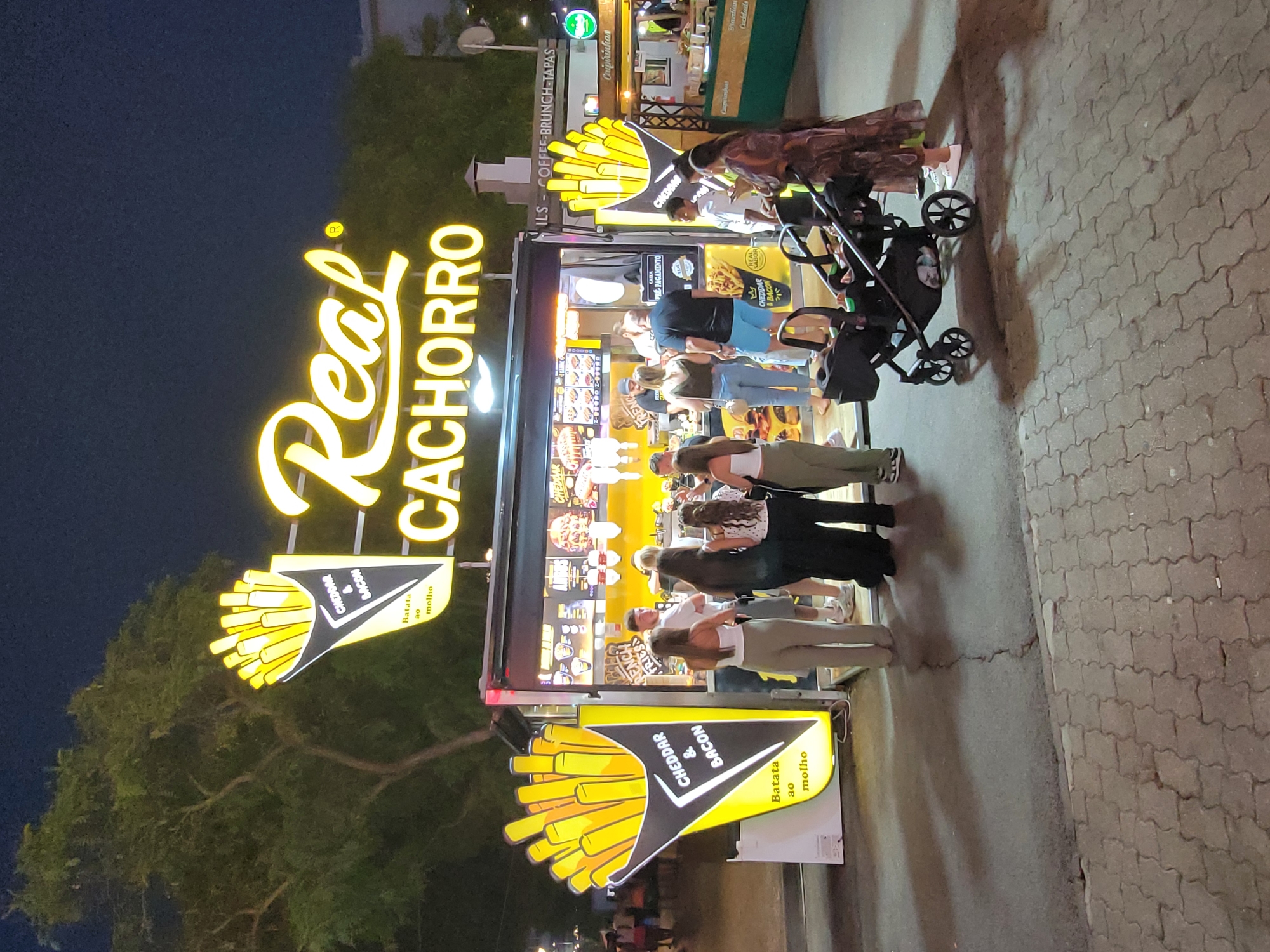 This is a vertical, slightly blurry nighttime photograph taken in Quarteira, Portugal. The primary subject is a vibrant food stand with a large, illuminated sign that reads "Real Cachorro" in bright yellow lettering. The stand appears to be a hot dog vendor, with images of french fries and toppings like cheddar and bacon displayed prominently on its exterior.
Several people are gathered around the food stand, either ordering or waiting for their food. Their faces are not clearly visible due to the angle and lighting, but they seem to be engaged in conversation or looking at the menu. A baby stroller is parked next to some of the patrons. The ground is paved with interlocking bricks.
In the background, other food stalls or establishments are visible, suggesting a lively market or boardwalk setting. One stall displays signs for "Caipirinhas," "Cocktails," and "Coffee-Brunch-Tapas," indicating a variety of food and drink options available in the area. The presence of trees on the left side of the image suggests an outdoor location. The overall atmosphere appears to be casual and bustling, typical of an evening gathering at a food market or tourist area. The time of day is clearly night, indicated by the artificial lighting and the dark sky.