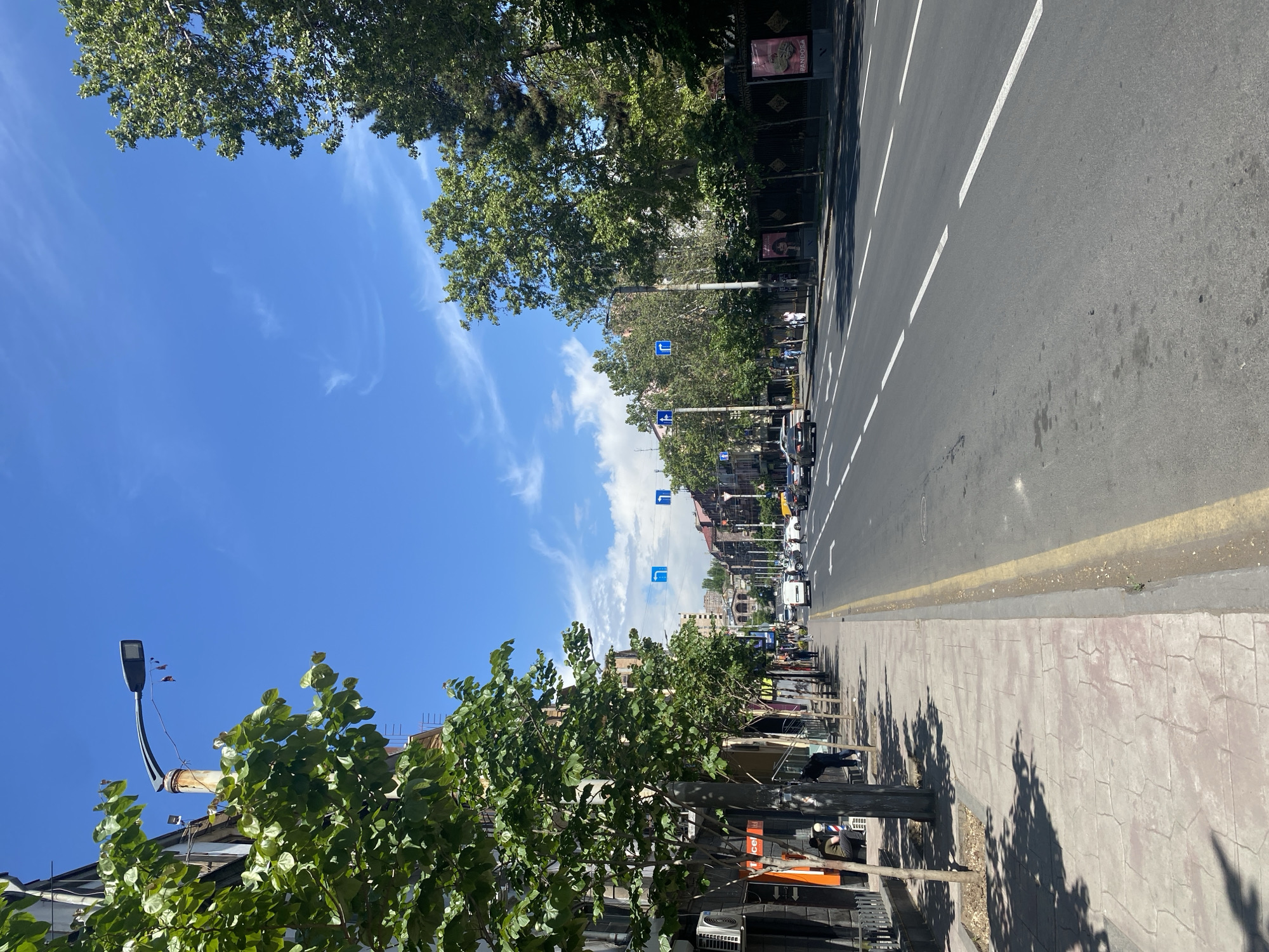 The image shows a street scene in Yerevan, Armenia on a sunny day. The view is down a wide city street with multiple lanes of traffic. Trees line both sides of the street, providing shade along the sidewalks. The street has white lane markings and a yellow line separating it from the sidewalk. There are parked and moving vehicles in the street. Traffic signs are visible periodically on the left-hand side of the road. People are walking along the sidewalks, some sitting near buildings on the lower left. Buildings are visible on both sides of the street. The sky is blue with scattered clouds.