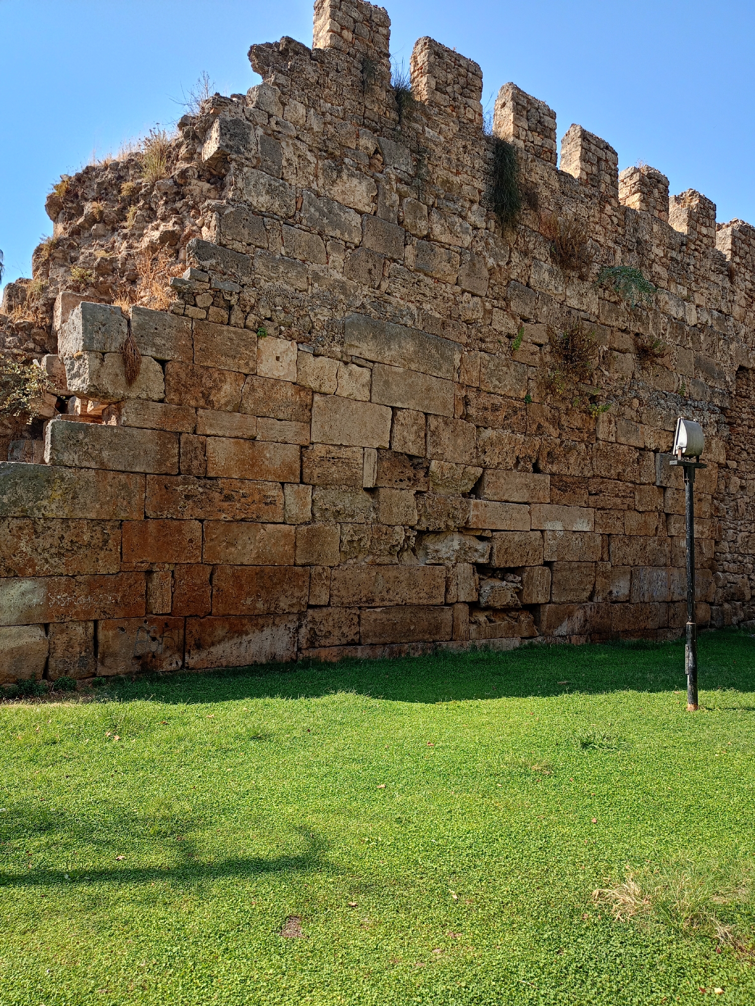 The image shows a section of ancient stone fortifications in Antalya, Turkey, under a clear blue sky. The wall, constructed from large, rough-hewn blocks of reddish-brown stone, exhibits signs of age and erosion, with some blocks chipped and missing. The top of the wall features crenellations, also made of stone. Sparse vegetation, including dry grasses and small green plants, grows in the crevices and along the top of the wall. In the foreground, a neatly trimmed, vibrant green lawn stretches out. To the right of the frame, a dark metal pole stands vertically, topped with a light fixture that appears to be a traditional lamppost with a metal shade. The sun casts shadows from the wall and the lamppost onto the lawn. The overall scene depicts a historical landmark integrated into a park-like setting.