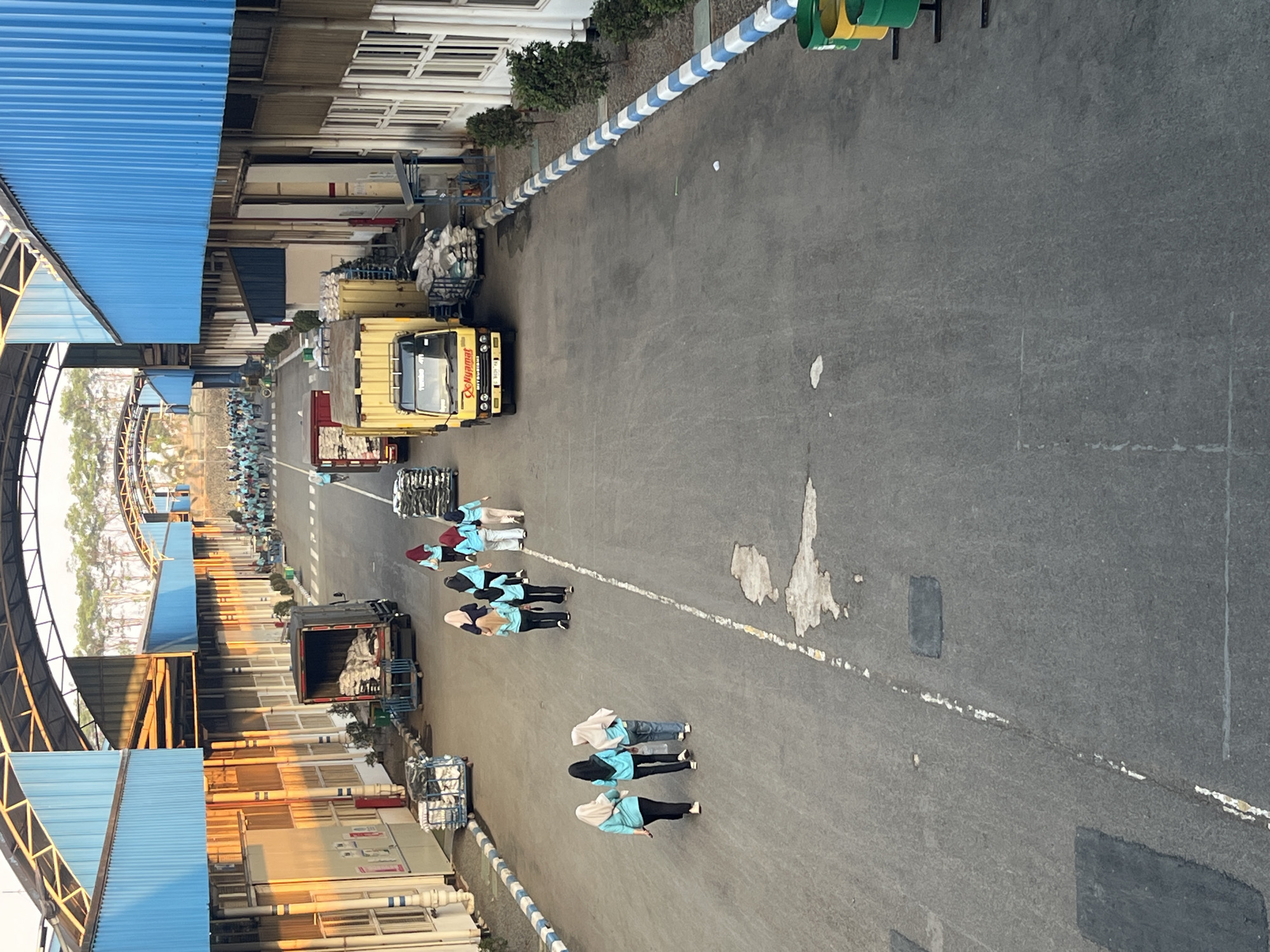 The image depicts an outdoor industrial area with buildings on either side of a wide paved road. The buildings feature blue corrugated metal roofs and lighter colored walls. On the left, a truck with an open bed filled with sacks is parked. Several people, mostly women wearing headscarves and light blue tops, are walking on the road. A yellow truck with "DNYamat" branding is also visible. In the distance, a large group of people in similar attire are seen walking towards the camera. The environment appears to be warm, with sunlight casting long shadows. The location is identified as an industrial complex in Pelang, Indonesia.
