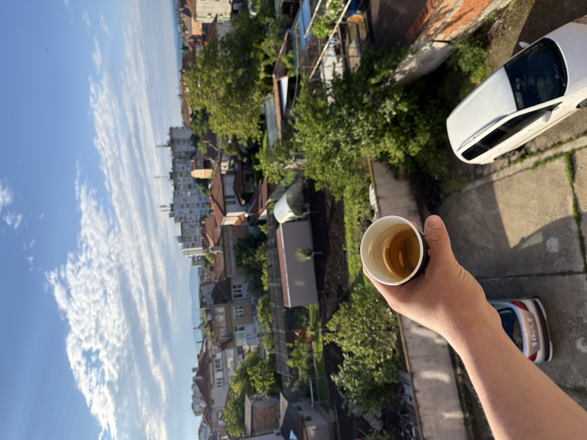 The image shows a person's hand holding a cup of coffee, with a view of Burgas, Bulgaria, in the background. The sky is blue with white clouds. Below, there are buildings, trees, and cars. Two cars are specifically visible: a white one and a silver one. The photograph appears to have been taken from a high vantage point.