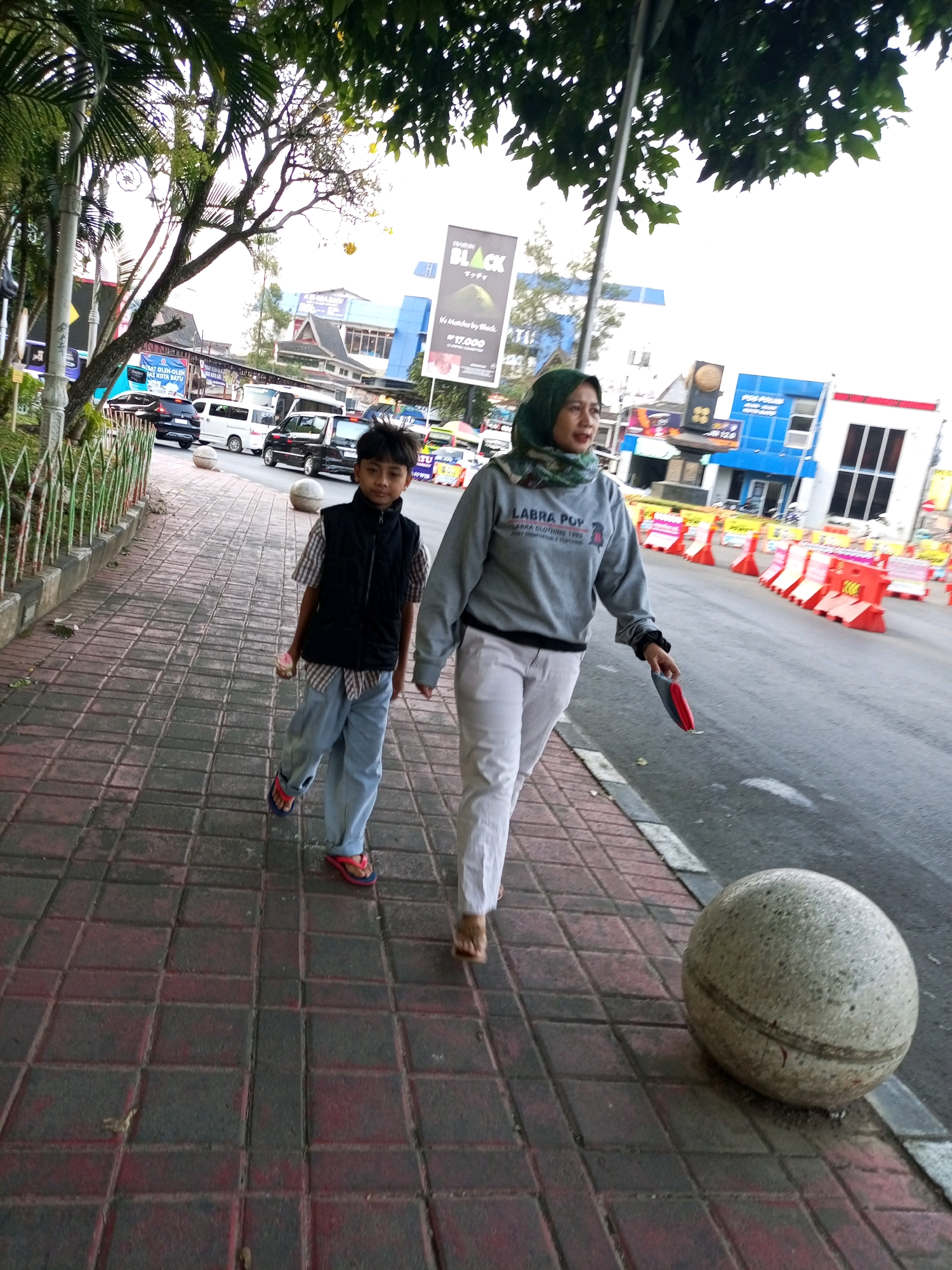 A street scene in Batu, Indonesia, features a sidewalk with red and gray square tiles. Two people are walking on the sidewalk. In the foreground on the left, a young boy wearing a plaid shirt, a black vest, light blue pants, and red flip-flops is walking. He is holding a small object, possibly a candy. To his right, a woman wearing a green hijab, a gray sweatshirt with the text "LABRA POP" on it, and white pants is walking. She is holding a red and blue object in her right hand. A large, round, speckled stone planter is situated on the edge of the sidewalk near the street. In the background, there are several vehicles, including black cars and white vans, and various buildings, some with blue facades. Traffic cones are visible along the side of the road. Overhead, trees with green leaves provide shade. A large billboard with the word "BLACK" is also visible.