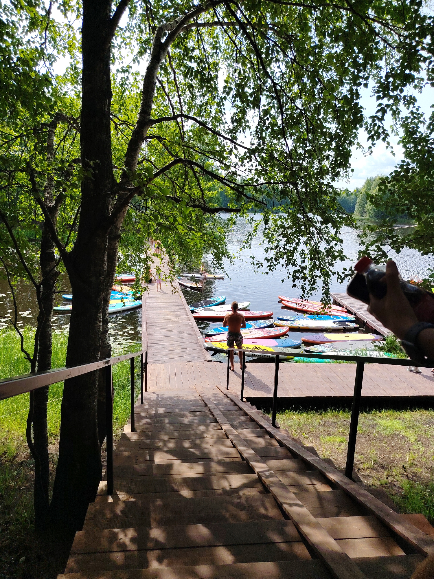 The image depicts a wooden staircase leading down to a wooden pier on a lake. The pier is lined with numerous colorful paddleboards. A shirtless man stands on the pier, facing away from the camera. In the background, other people are visible on the pier and in the distance. Trees with green foliage overhang the scene, casting shadows on the stairs and pier. The lake appears calm and reflects the sky. The location is identified as the village of Vylytdor, Russia.
