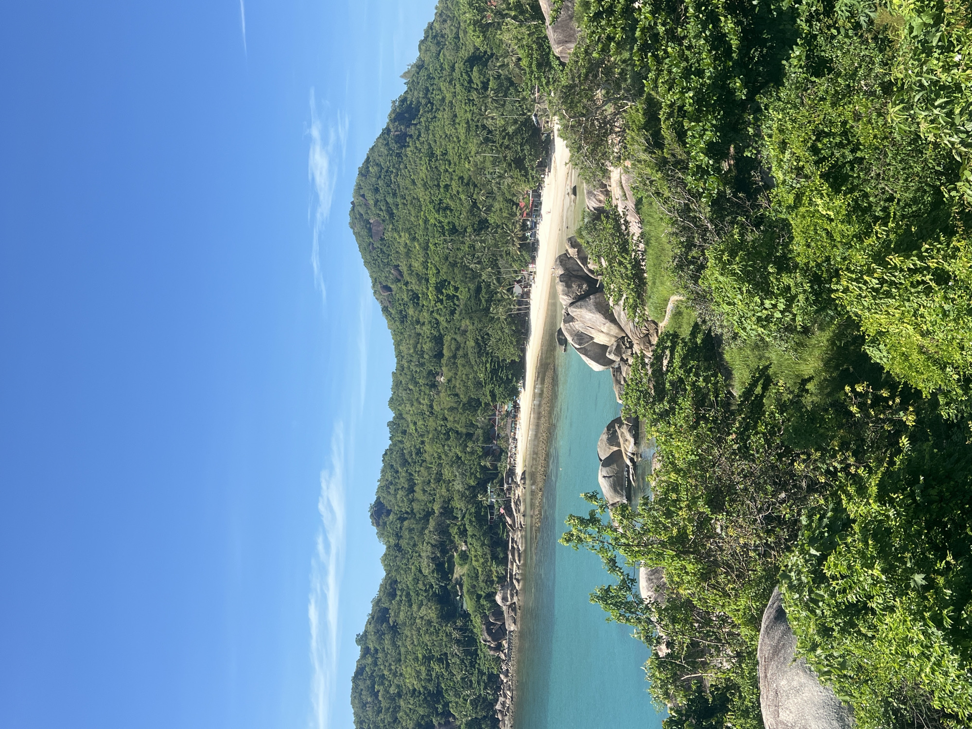 An aerial view captures a tropical beach scene under a bright blue sky. Lush green hills covered in dense foliage and palm trees form a backdrop to a crescent-shaped white sand beach. The turquoise water is calm and shallow near the shore, with some large grey rocks dotting the coastline and partially submerged in the water. A few small buildings, possibly bungalows or shops, are visible along the edge of the beach, nestled amongst the trees. The foreground is dominated by thick green vegetation and large boulders, suggesting a slightly elevated vantage point overlooking the bay. There are no visible people or active events in the scene. The location is identified as Baan Chaweng Noi, Thailand.