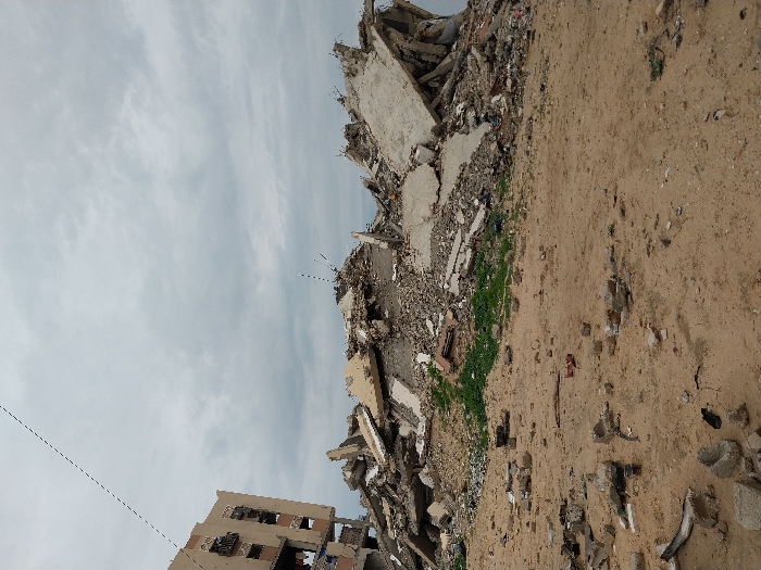 Extensive debris and rubble dominate the scene, likely the remnants of destroyed buildings. Some vegetation sprouts along the edges of the wreckage. A partly demolished multi-story building remains in the foreground. A sandy ground with debris scattered across it stretches out, contrasting the sky filled with scattered clouds.