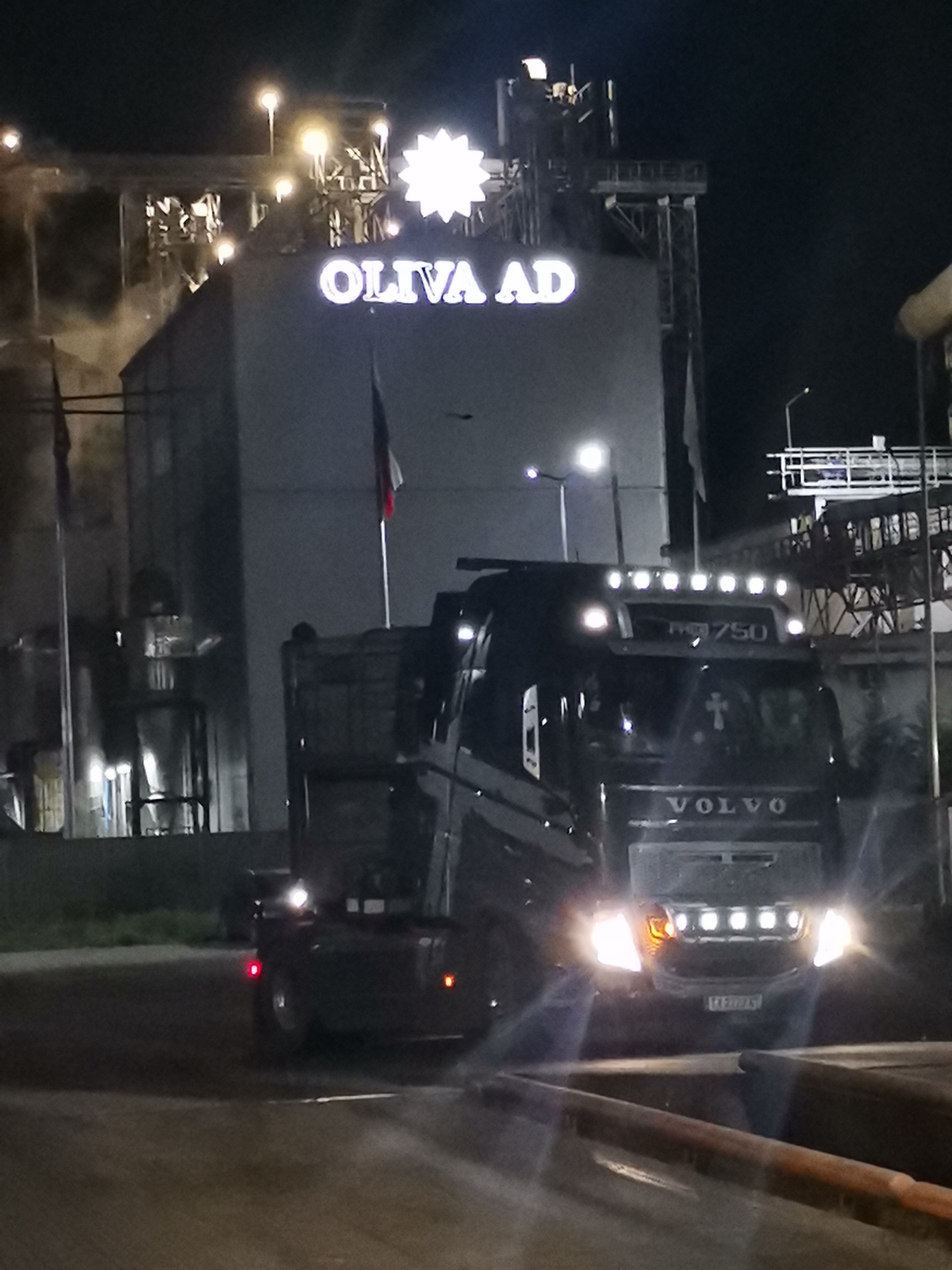 The image depicts a nighttime scene in Razdelna, Bulgaria, showing an industrial complex with a large building prominently displaying "OLIVA AD" in illuminated lettering. A bright, star-shaped symbol is mounted above the name. A Volvo truck with its headlights on is positioned in front of the building. The truck also has additional lights on its roof. The surrounding area appears to be a paved industrial yard with flags. Smoke is rising from the industrial complex.