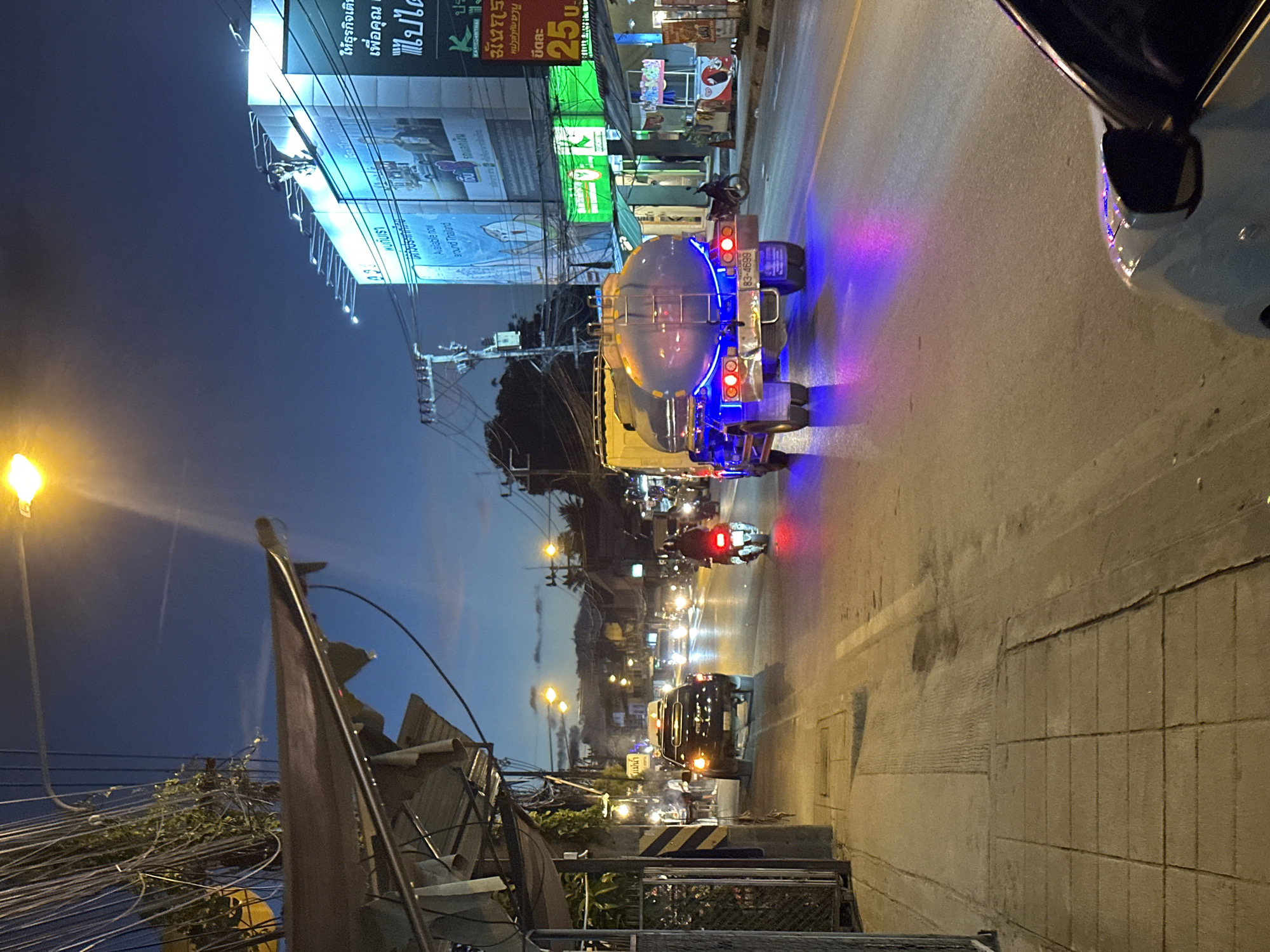 This media file captures a nighttime scene on a street in Baan Mae Nam, Thailand. The primary focus is a white tanker truck with a blue tank, illuminated by vibrant blue and red lights as it drives down the road. Several motorcycles are also visible, with one appearing to be following closely behind the tanker. The street is lined with buildings and illuminated by streetlights and the glow from shop signs, suggesting a commercial or residential area.
Above the street, a large billboard displays text in Thai, including what appears to be an advertisement. The sky is dark, indicating it is nighttime. The overall atmosphere is that of a bustling street scene at night, with the glowing lights of vehicles and signs creating a dynamic and colorful environment.
Visible text includes "แม่น่า", "83-4699", "พบกับเรา", "Available now", "sound Thailand", "รถแลกเงิน", "ให้ธุรกิจเติธ", "เพื่อคุณ", "ไปได", "KAVISSANTHAL", "ปร", "มันাছে", "หม่าส่งทัษษาน", "ขีดละ", and "25 U". The presence of these signs and the traffic suggest normal activity in a Thai town or city after dark.