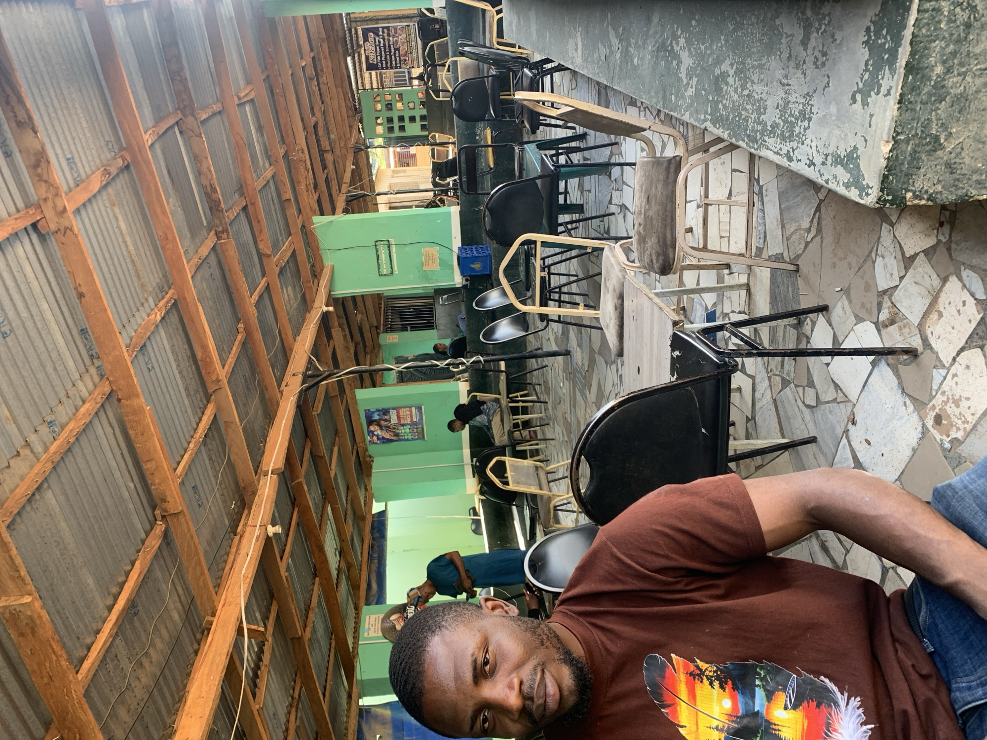 This image depicts a semi-enclosed, casual eatery or gathering space located in New Stadium, Nigeria. The scene is illuminated by natural light, casting distinct shadows. In the immediate foreground and to the left, an adult male with a dark complexion, short dark hair, and a trimmed beard is seated, facing directly towards the camera with a neutral expression. He is wearing a dark brown t-shirt featuring a stylized feather graphic on the left side, which depicts a sunset over a body of water with silhouetted trees and a small boat. He is also wearing blue denim jeans. His left forearm is visible, resting on his lap or an unseen surface. In the mid-ground, to the left of the main subject, another adult male with a dark complexion is standing, partially facing away from the camera. He wears a dark blue tunic and a dark cap, and appears to be looking down at a small dark object held in his hands. Further back, another adult male with a dark complexion is seated at a table, facing mostly away from the camera, wearing a light-colored long-sleeved shirt and dark trousers. A fourth, indistinct figure, possibly female, is seated in the far left background. No direct interactions between individuals are discernible. The environment features a ceiling constructed from corrugated galvanized iron sheets, supported by a framework of rough-hewn wooden beams and planks. Exposed electrical wiring and a single bare light bulb are visible hanging from the wooden structure. The interior walls and structural pillars are painted a light green, with some areas showing signs of wear, chipping, and dirt. On a green wall in the mid-ground, a poster for "Obiagu Lager Beer" with text about pricing is affixed. On a wall to the right, a sign titled "Faith Kitchen" lists a menu of food items including "Cat Fish Pepper Soup" and "Plantain & Scrambled Eggs." The floor is a mosaic of irregularly shaped, light-colored tiles (off-white, grey, light brown) with visible grout lines and areas of discoloration. The space is furnished with multiple chairs and small tables. The chairs vary in design; many are black metal chairs with black seats, while others are light-colored metal chairs with worn, light-colored seats and backs. One light green chair is also present. The tables are simple, square or rectangular, with light-colored, worn surfaces. A blue plastic crate is visible on the floor near the center-left. A