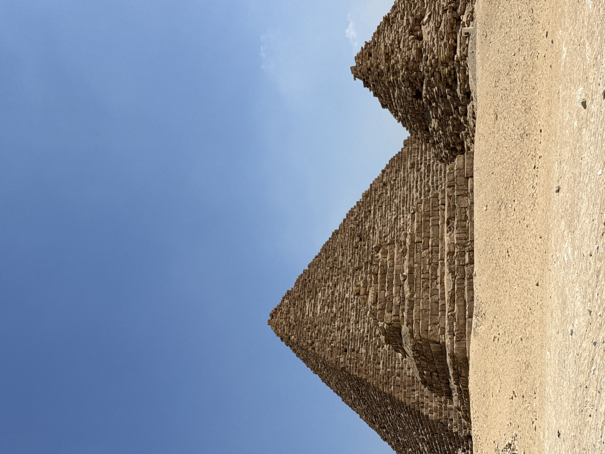 The image shows two ancient Egyptian pyramids against a clear blue sky. The larger pyramid on the left is the Great Pyramid of Giza, also known as the Pyramid of Khufu. Its stone blocks are visible, and the structure appears weathered. To the right of the Great Pyramid is a smaller, less intact pyramid, possibly the Pyramid of Khafre or Menkaure. The foreground consists of a sandy desert landscape with small stones and pebbles. In the far right, a small, white modern structure is visible near the base of the smaller pyramid. No people are present in the image.