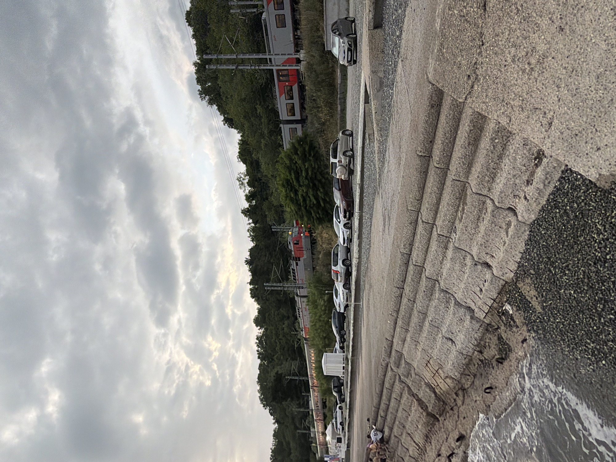 The image depicts a coastal scene in Sochi, Russia, with a train traveling along tracks elevated above a parking lot filled with cars. The foreground features a wave-lapped concrete embankment leading to a rocky shore. Several people are seated on the embankment, facing the water. Behind the embankment, a gravel parking lot hosts a variety of vehicles, including sedans, SUVs, and a small hatchback. Further back, a red and gray passenger train is visible, moving from left to right. The train is flanked by lush green trees and dense foliage on a hillside. Overhead, a cloudy sky with patches of diffused sunlight contributes to the overcast atmosphere. The overall impression is a blend of industrial infrastructure and natural coastal beauty.