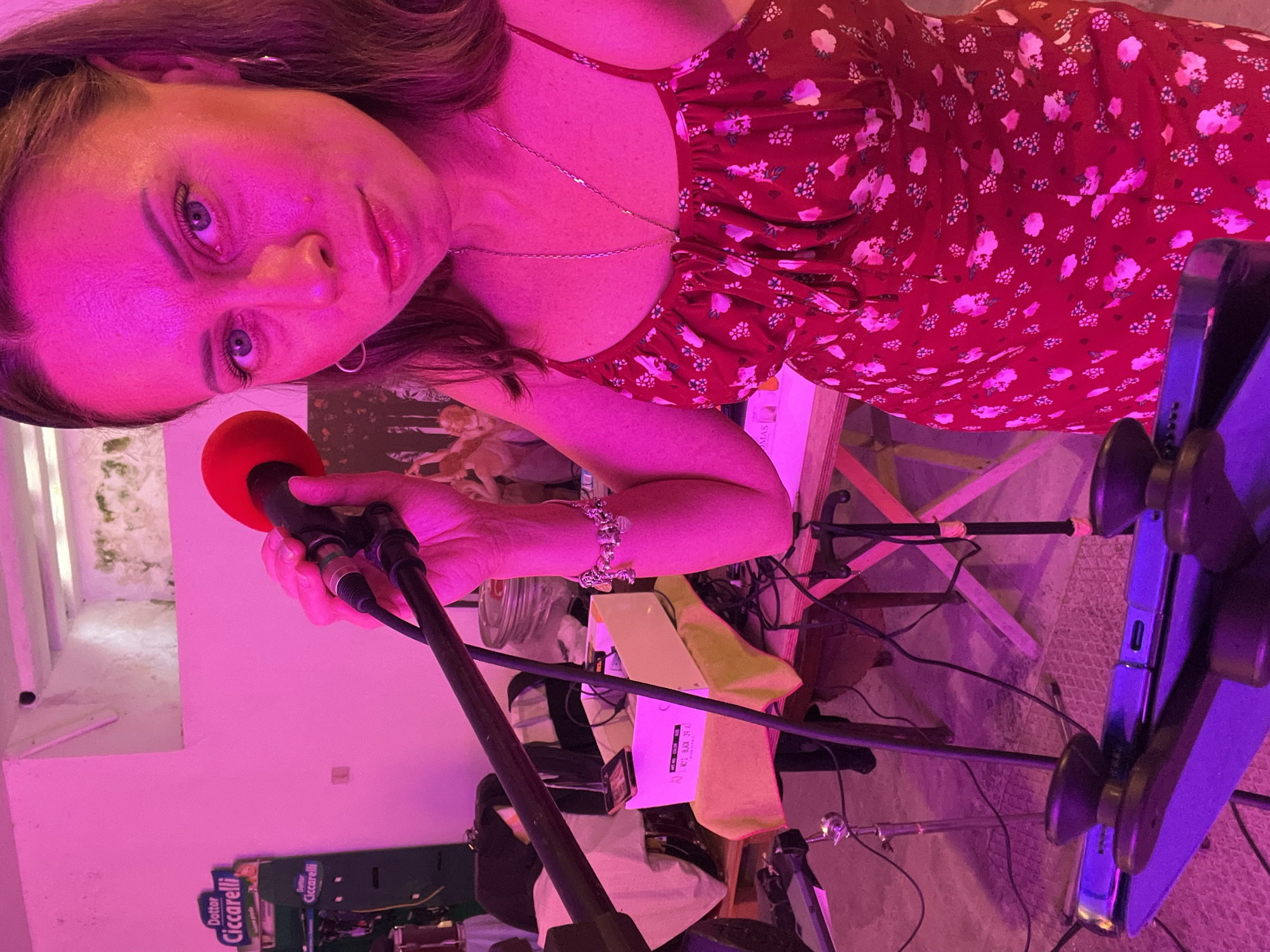 A young adult female is depicted holding a microphone with a red foam windscreen in her right hand, positioned as if performing or speaking, under intense magenta lighting in an indoor setting in Santeramo in Colle, Italy. She has dark hair, blue eyes, and is wearing a red floral sundress with thin straps, a silver necklace, silver hoop earrings, and a silver charm bracelet on her right wrist. Her expression is neutral as she looks directly at the camera.
Her right hand grips the black microphone, which is connected to a cable extending downwards alongside a black microphone stand. In the immediate foreground, a dark music stand or tablet holder is visible, displaying a dark, reflective surface, likely an electronic device.
The background is characterized by a cluttered, semi-industrial or event space. To the left, a green "Dottor Ciccarelli" advertising sign is prominent, positioned above another similar sign. A maroon drum, likely a tom, rests on a white cloth-draped surface, supported by a wooden pallet or low platform. Various cables and other dark equipment are scattered on this platform.
Behind the woman, a faded wall mural or tapestry depicting classical figures is discernible, heavily tinted by the magenta light. To her right, a wooden table holds a white cardboard box (partially labeled with text including "BLACK 29.5"), a black smartphone, and a clear glass jar with a red lid. Exposed cables, additional stands, and wooden table supports are visible throughout the midground. An architectural cut-out or alcove in the upper wall reveals an older, discolored wall texture, possibly stone or concrete with some growth. The floor appears to be a light-colored, textured surface, also saturated in pink and purple hues.
