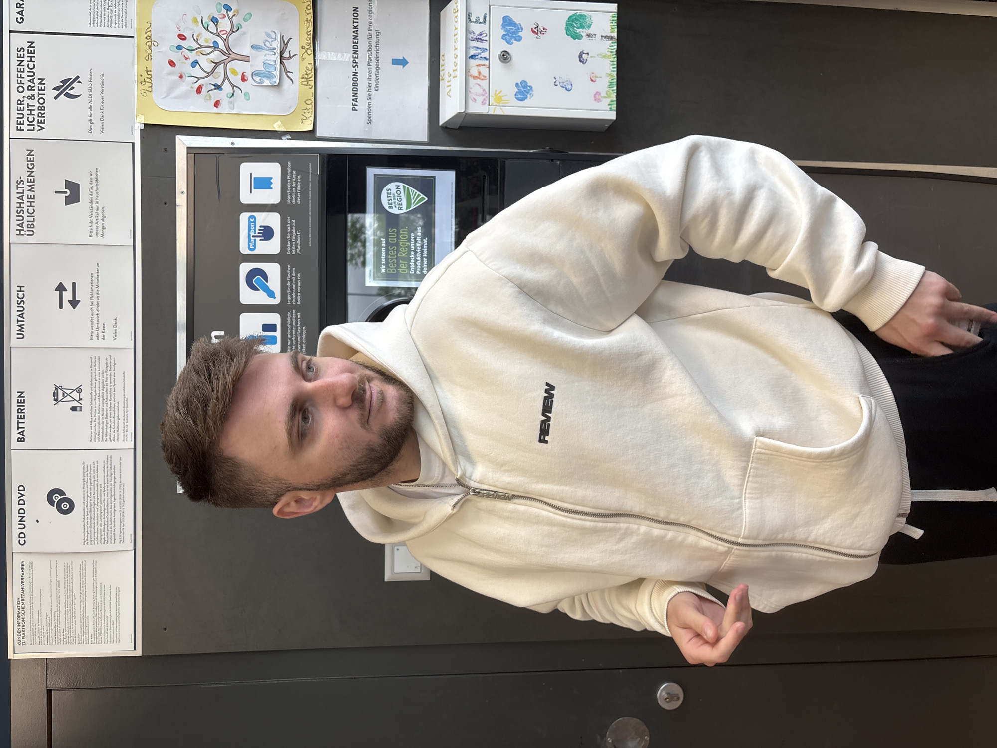 A person stands beside an automated bottle return machine in Kaarst, Germany. The machine is located on a dark gray exterior wall, next to multiple informational signs: one about electronic payment methods, followed by information about CD/DVD, batteries, product returns, household quantities, a fire safety notice, a thank-you note from "Kita," another sign about bottle donations to local childcare. To the right of the signs, a donation box is mounted on the wall. The person wears a cream-colored "REVIEW" hoodie with the zipper partially undone and gives the middle finger with his right hand.
