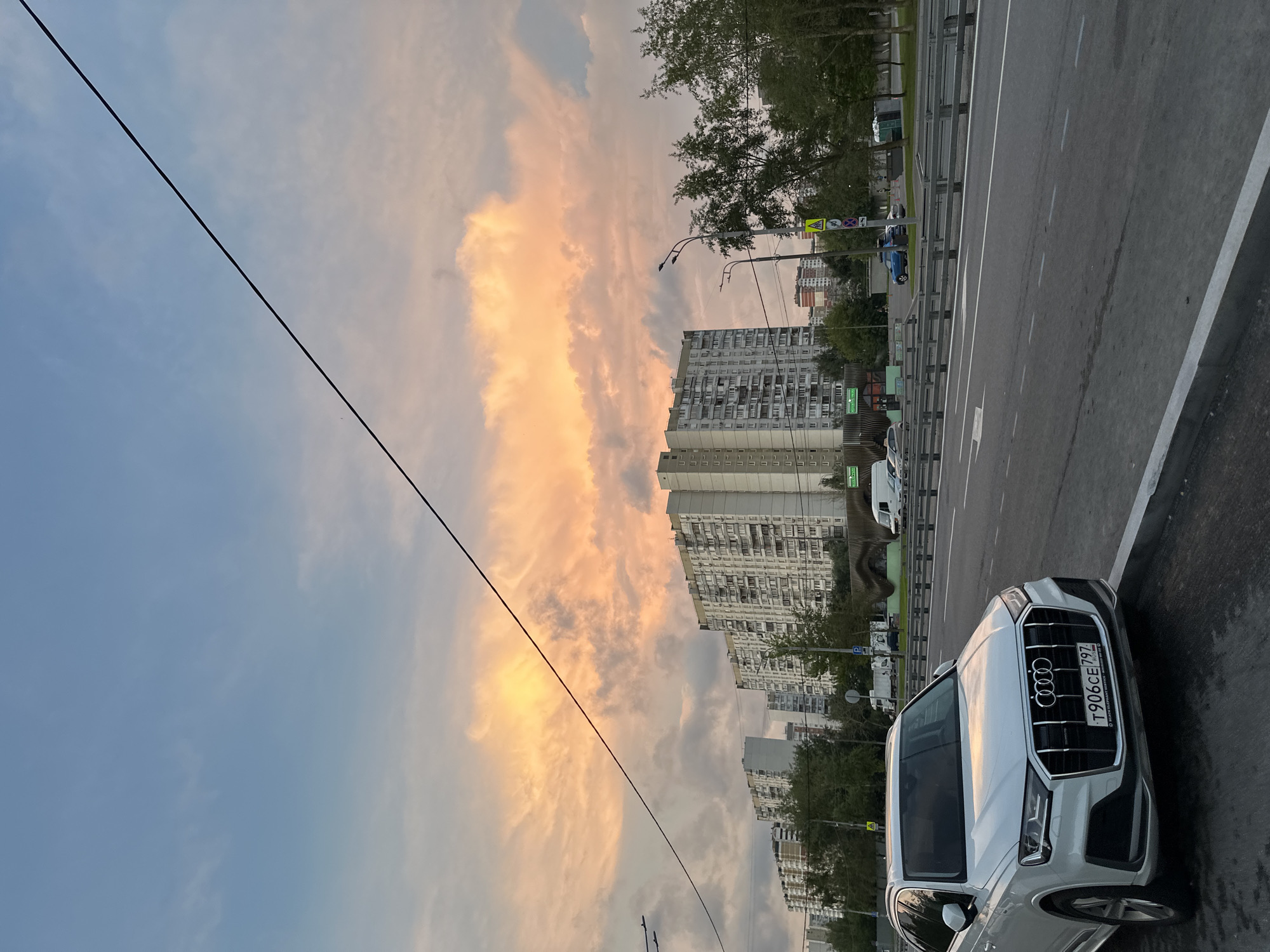 The image depicts a white Audi Q7 parked on the side of a road in Moscow, Russia, facing the camera. The car's license plate reads "T906CE 797". In the background, several large apartment buildings are visible, typical of Soviet-era architecture. The sky is filled with dramatic clouds, tinged with orange and pink hues, suggesting either sunrise or sunset. Trees line the road, and a street light stands near the car. The overall scene appears to be an urban landscape during twilight.