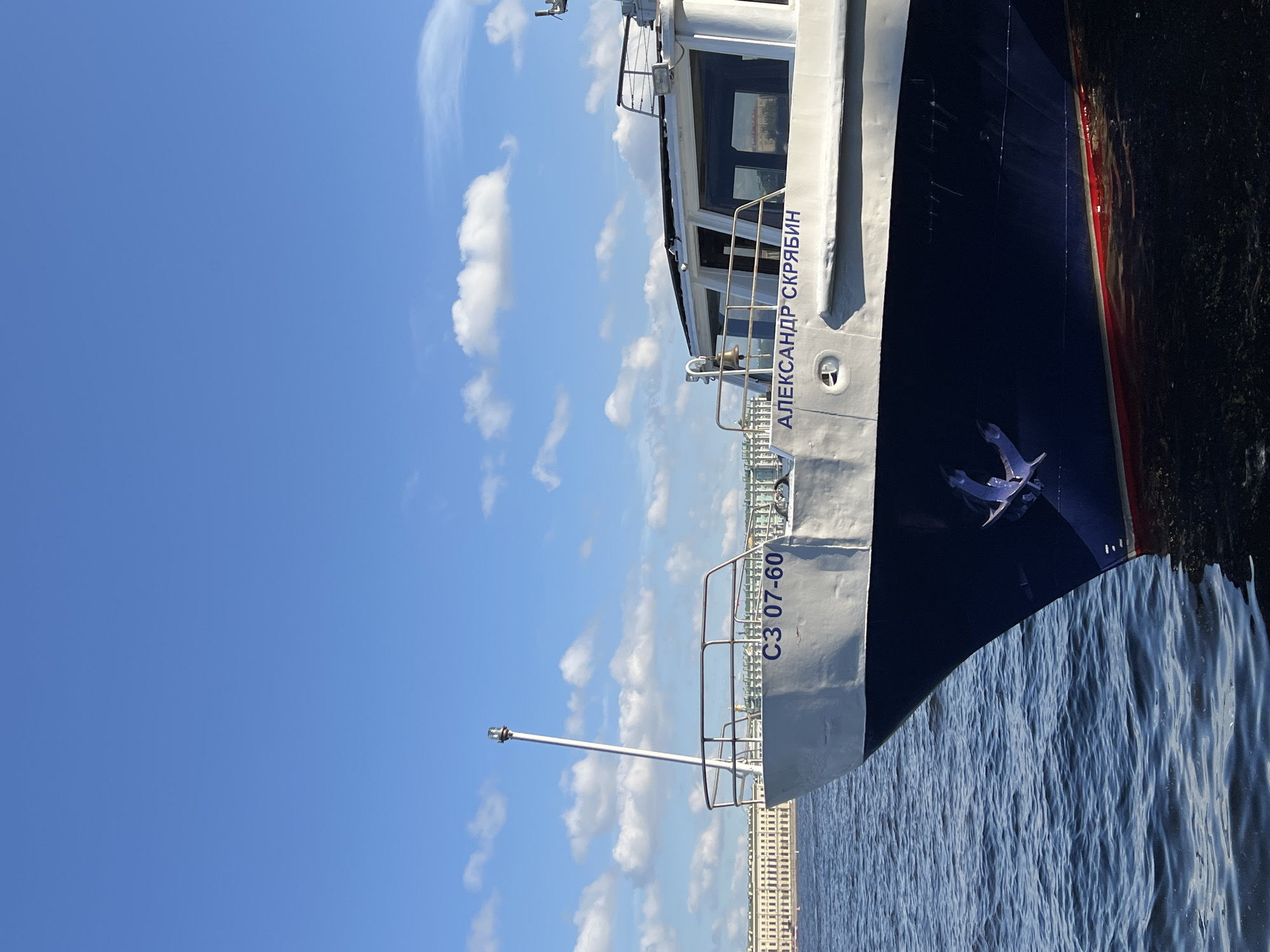 The image shows a segment of the river cruise ship "Aleksandr Scriabin" in Saint Petersburg, Russia. The white and blue ship is partially visible, featuring its name in Cyrillic script, the registration number "C3 07-60", an anchor, and a white signal mast. The ship is situated on the Neva River, with ripples on the water visible. The sky is blue with scattered white clouds. A building is seen on the bank of the river.