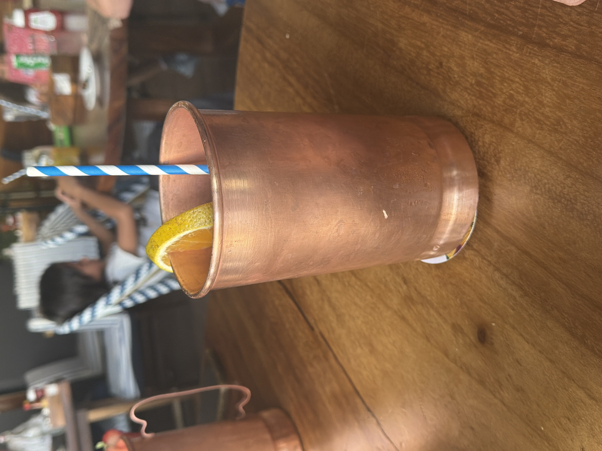 The image shows a copper cup containing a drink with an orange slice and a blue-and-white striped straw. The cup rests on a wooden table with visible grain. The background features blurred elements, including what appears to be a person seated on a blue-and-white striped chair and other indistinct objects on a table behind the cup. The scene is likely a restaurant or bar setting. The information about Puebla, Mexico is not discernible from the image.
