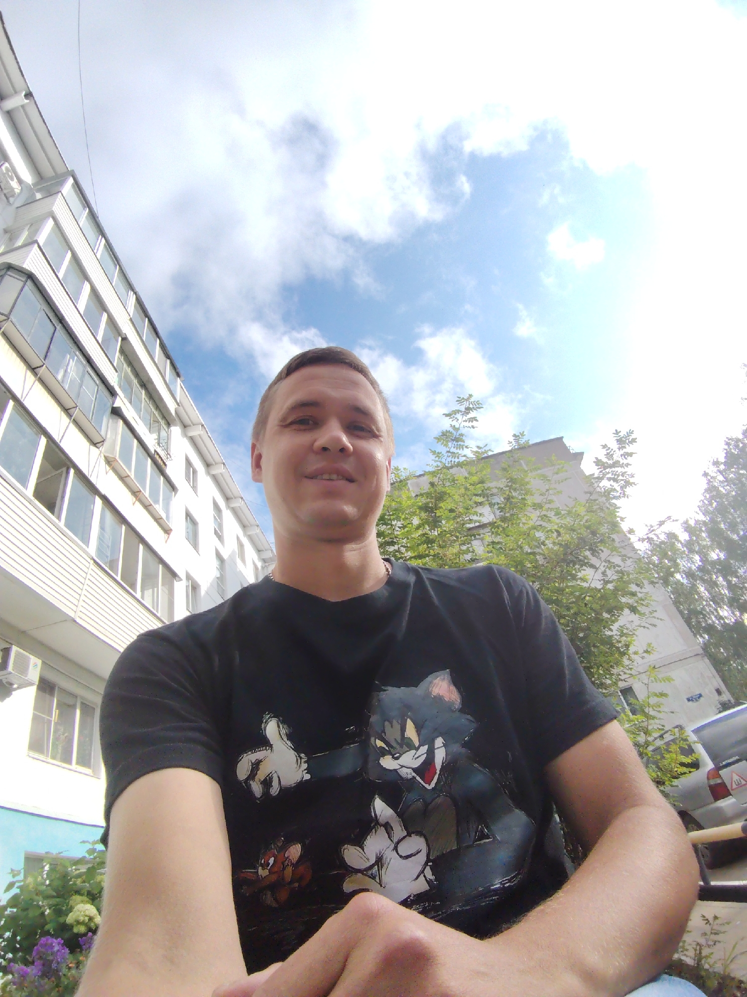 A medium-shot, outdoor, eye-level, wide-angle selfie shows a man with short, light brown hair and a fair complexion. He is smiling at the camera, wearing a black t-shirt with a graphic print of Tom and Jerry characters. His left arm is in the foreground, resting on his lap.
Behind the man, on the left, is a multi-story apartment building with white siding and rows of windows. An air conditioning unit is visible on the lower left portion of the building. To the right of the building, a large tree with green leaves is prominent. Further in the background, another apartment building is visible. The sky is blue with scattered white clouds.
The scene appears to be in a residential area, likely in the city of Chaikovsky, Russia, based on the architectural style of the buildings and the surrounding vegetation. The lighting suggests it is daytime, possibly late morning or early afternoon, given the angle of the sun and the shadows. The presence of a parked car in the background on the right suggests a street or parking area adjacent to the residential buildings.