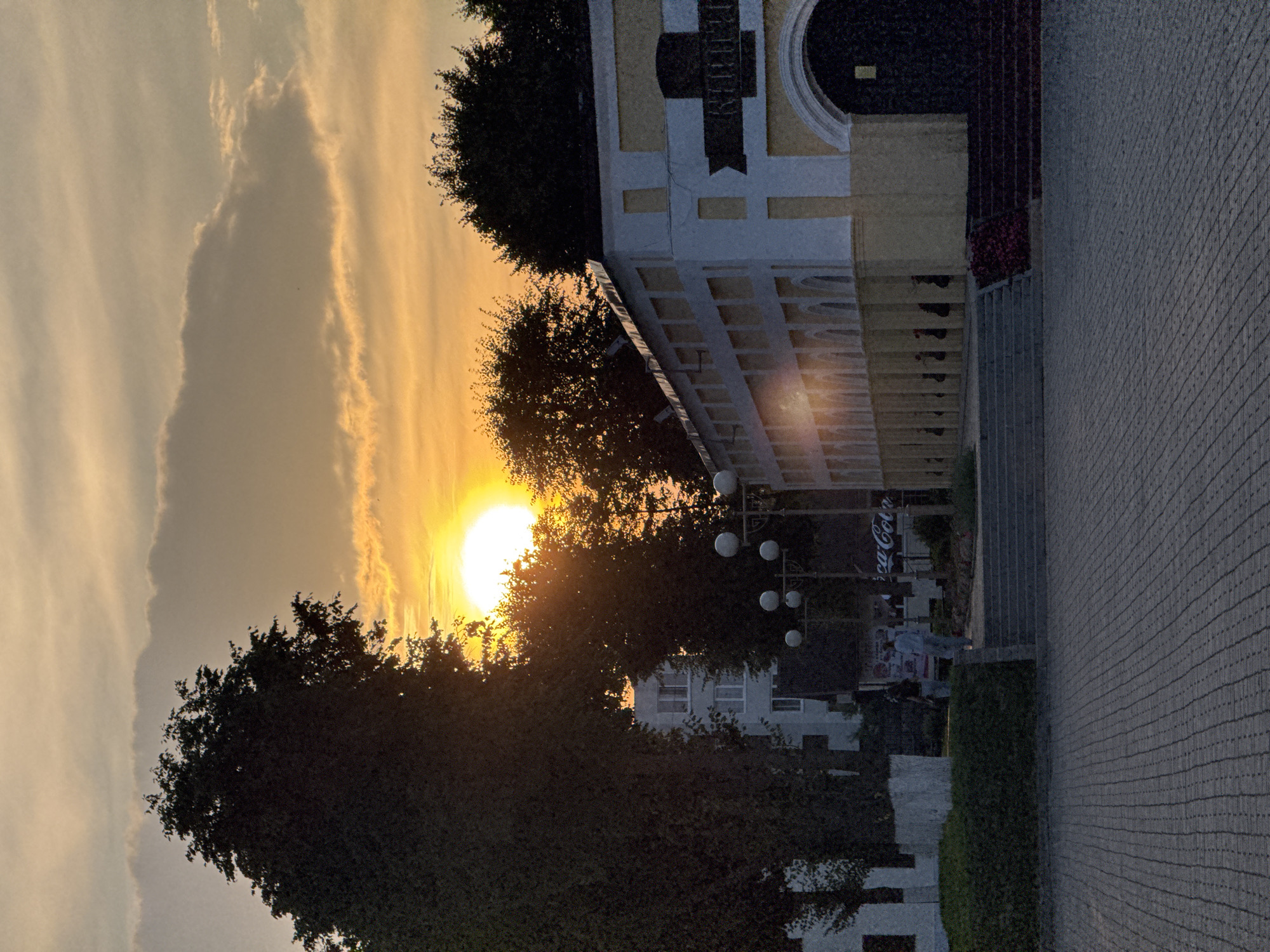 The image depicts a sunset scene in Niasvizh, Belarus. The sun, a bright orange orb, is low in the sky, casting a warm glow and creating a silhouette of trees. In the foreground, a paved area transitions to a raised walkway with steps. To the right, a building with a yellow and white facade and arched entrances stands prominently. A dark banner with lettering is affixed to the building. To the left, lush green bushes and trees line the walkway. In the background, a multi-story building is visible behind the trees, and a Coca-Cola advertisement is partially visible. Two individuals are seen walking on the raised walkway. Streetlights with multiple spherical lamps are also present. The sky is filled with streaks of orange and yellow clouds.