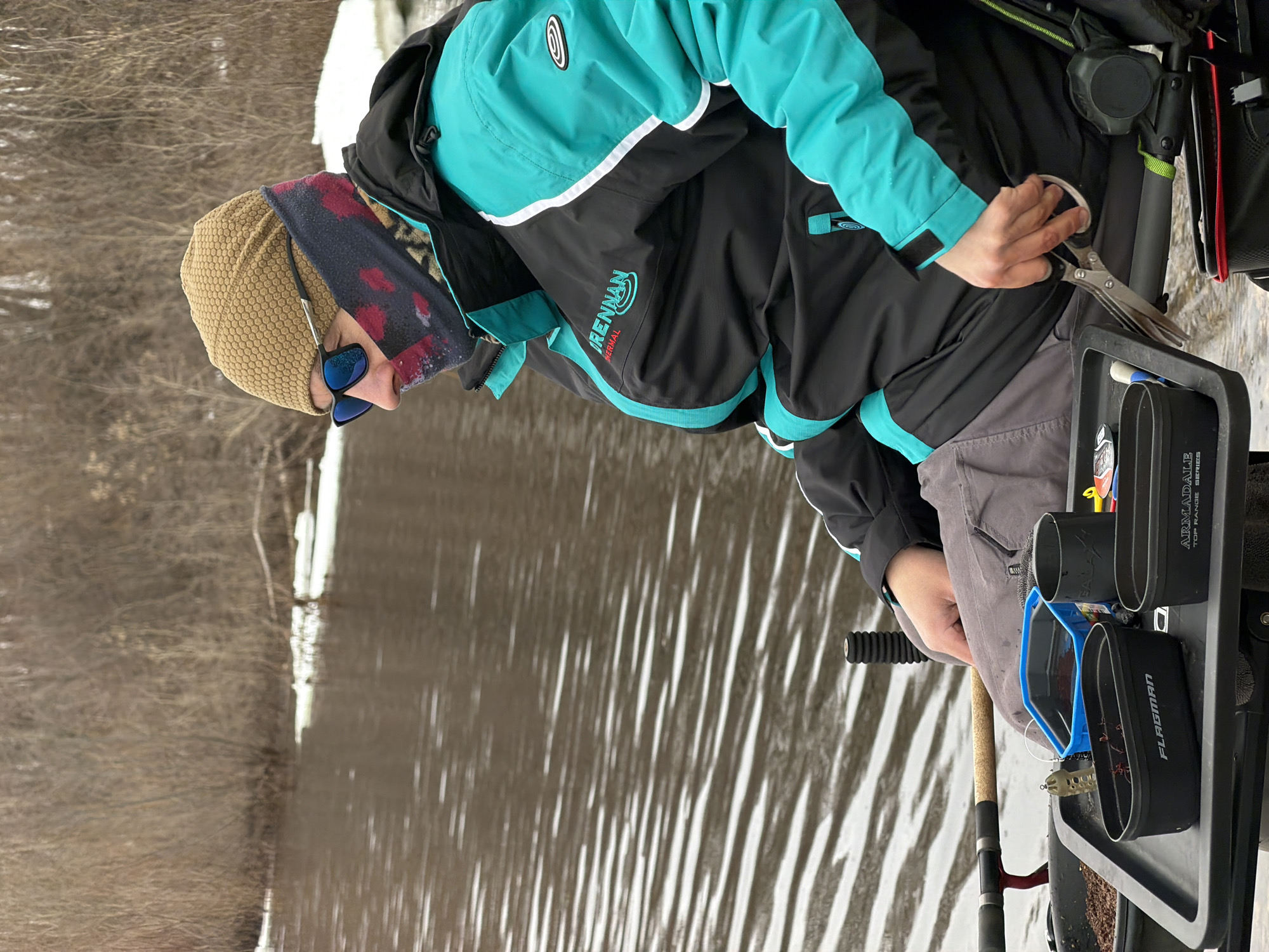 A person wearing a tan beanie, sunglasses, and a black and turquoise jacket with the word "DRENNAN" on the sleeve is shown in profile view, near a body of water with reflections of trees and sky. They are holding scissors near a tray with fishing supplies, including bait containers labeled "ARMADALE" and "FLAGMAN." A fishing rod is visible beside the tray. The location appears to be outdoors, possibly near a river or lake in a natural environment. There is evidence that the image may be associated with Chaikovsky, Russia.