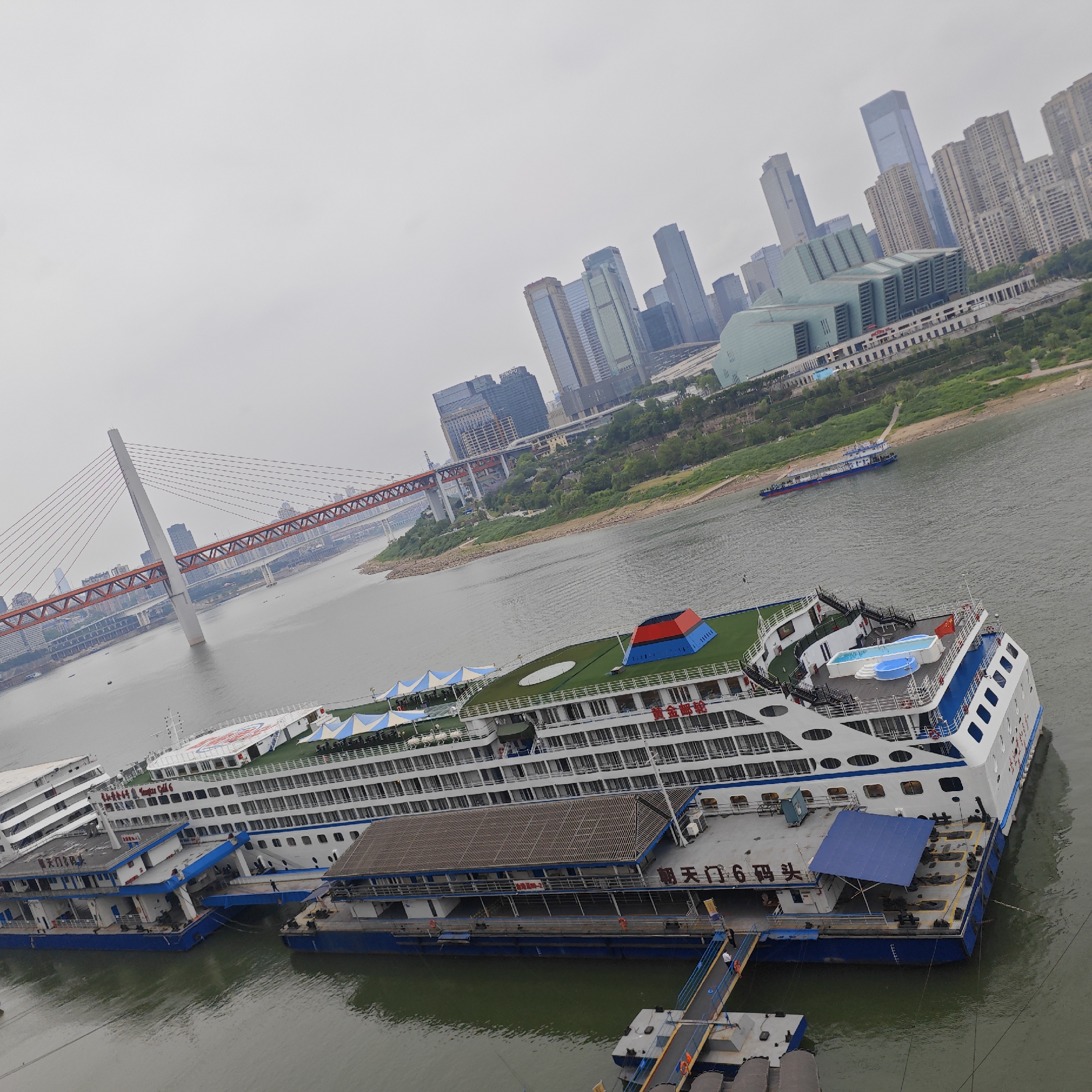 The image depicts a cityscape in Yuzhong District, China, featuring a river with several large cruise ships docked along its bank. The ships are primarily white and blue, with multiple decks and various amenities visible, such as seating areas and small pools.
In the background, a large red suspension bridge stretches across the river. Further behind, a skyline of modern skyscrapers and buildings rises into the hazy, overcast sky. The river is calm, with a slightly greenish hue, and a smaller boat is seen moving on the water's surface. A pedestrian walkway leads to the docked ships, suggesting active passenger traffic. The overall scene conveys a sense of urban activity and transportation in a densely built environment.