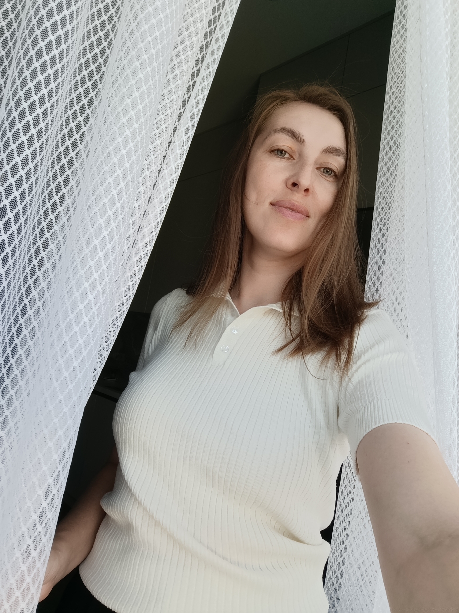 A woman is standing indoors, partially obscured by a white, lace-like curtain. She wears a short-sleeved, ribbed white polo shirt. Her hair is brown and shoulder-length. The background is dimly lit, with indistinct objects suggesting an interior space. Based on the context of being in Chaikovsky, Russia, the location is likely an apartment or home setting in that city.