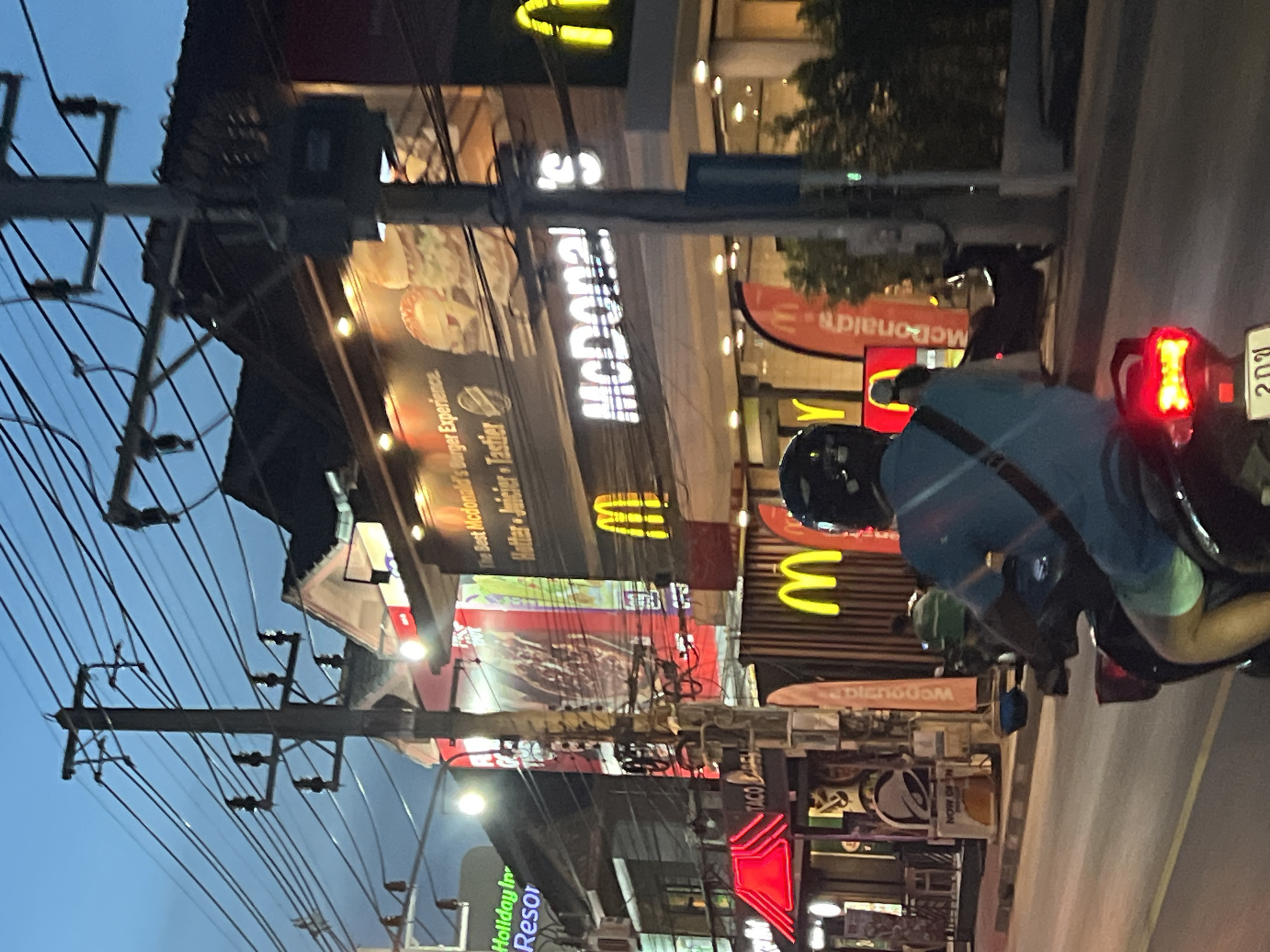 An eye-level, medium shot captures a street scene in Baan Bo Phut, Thailand, at dusk. The foreground is dominated by the rear of a black scooter, with a rider wearing a light blue t-shirt and shorts, and a black helmet. The scooter's red taillight is illuminated. The street is paved with asphalt and marked with yellow lines.
In the mid-ground, to the left, a sign for "Holiday Inn Resort" glows in neon green. Adjacent to it, a bright red awning stretches over a storefront, with a "TACO BELL" sign visible beneath. A "NOW OPEN" sign is also present. Further right, a McDonald's restaurant is prominently displayed with its signature golden arches. A large banner with a McDonald's logo is also visible. Two yellow McDonald's 'M' logos are also visible on the building.
The background features a network of utility poles and tangled electrical wires against a darkening blue sky. The architecture of the buildings suggests a commercial area, with multiple stories and illuminated storefronts. The overall impression is a bustling urban environment illuminated by artificial light as night approaches.