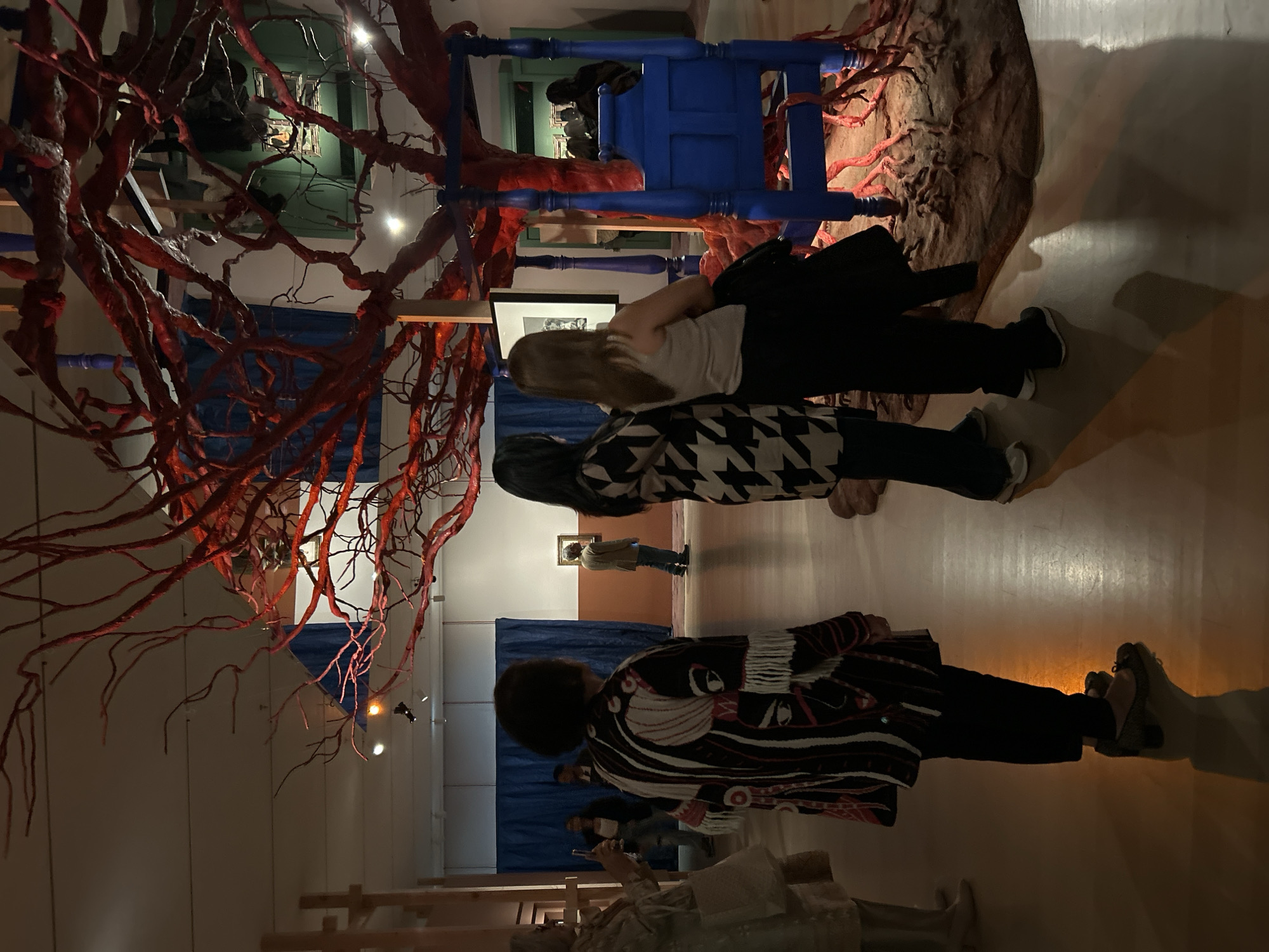 A horizontal, indoor shot shows an art installation, which may be in New York City, given the request. There are four visible people standing on a polished wooden floor. At the front, a person with short dark hair, wearing a long black-and-white patterned jacket, is looking at the art, while a woman with blonde hair is next to them, looking at a piece of art hanging on the wall behind them. Further behind is a man wearing a tan shirt and blue pants looking at another piece of art hanging on the wall behind him. In the front left is part of a nude statue and what appears to be a ladder.
The art installation includes a bright blue chair-shaped structure, with a large, red, root-like sculpture, possibly made of clay or plaster, wrapping around it. Behind the blue structure, one section has a blue tarp hanging and also a black and white drawing hanging on the wall behind the blonde woman.