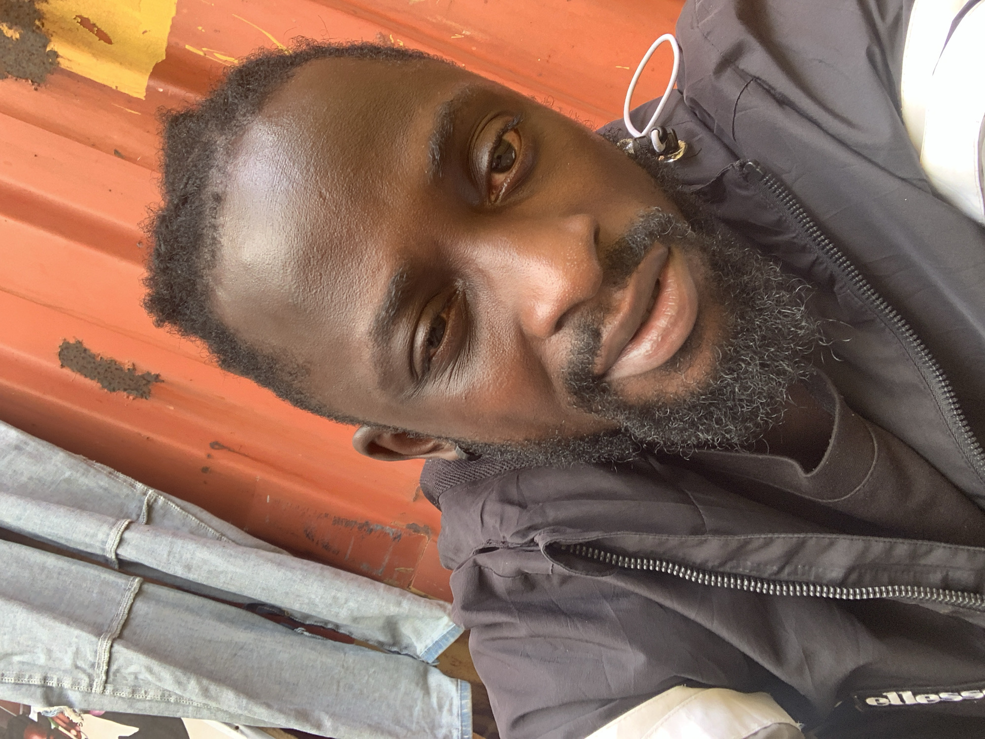 A dark-skinned man is featured in a close-up, angled selfie, looking directly at the camera with a calm, slightly smiling expression. He has short, dark, curly hair and a short, dark beard. His forehead exhibits a subtle sheen.
He is dressed in a dark (likely black or dark grey) zippered jacket, worn over a black t-shirt. A white loop or cord is visible near the jacket's collar, and the brand name "ellesse" is partially visible in white text on the lower right portion of the jacket.
The background is a prominent orange, corrugated metal surface, likely a wall or a shipping container, with some areas showing faded yellow paint or splatters. In the bottom left, folded light-colored denim trousers or jeans are visible.
The scene appears to be set in a casual indoor or enclosed environment, possibly a market stall, storage area, or workshop, consistent with a setting in Lekki, Nigeria. The lighting suggests it is daytime. The man's overall demeanor is relaxed.