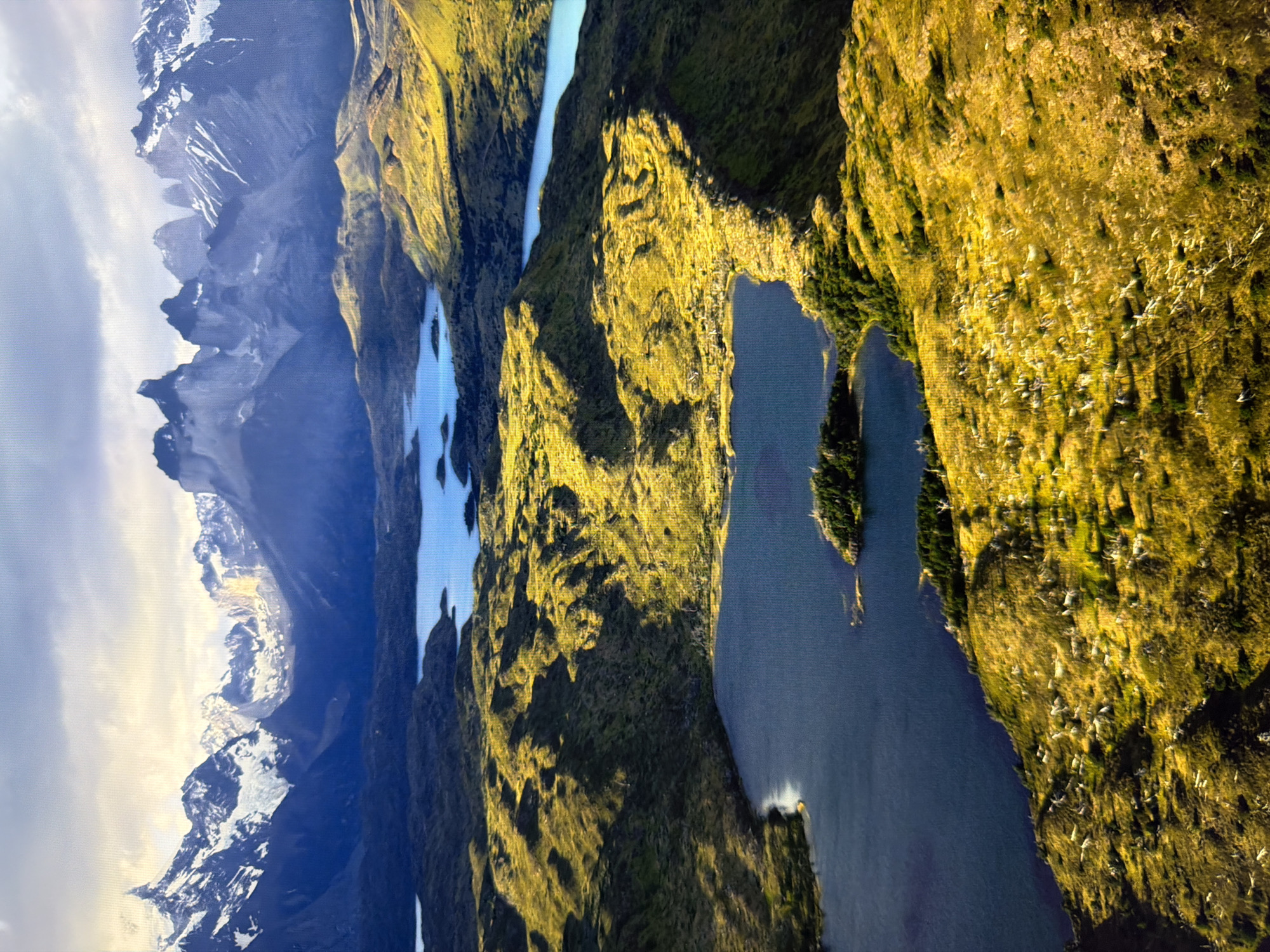 This is a high-angle aerial photograph of a mountainous landscape with lakes. In the foreground, a dark blue lake occupies the lower left quadrant, with a small, tree-covered island near its center. The surrounding terrain is characterized by rolling hills covered in golden-yellow grass, interspersed with patches of darker green vegetation and white debris. Shadows are cast across the hills, indicating the direction of sunlight. Further into the mid-ground, other bodies of light blue water are visible, nestled among more hills. The background is dominated by a range of majestic, snow-capped mountains with jagged peaks. The sky above is partly cloudy, with sunlight breaking through, illuminating parts of the mountains and hills. There are no people, animals, or man-made structures immediately discernible in the image, suggesting a natural, remote wilderness. The image does not depict the city of Kyiv, Ukraine; it shows a natural landscape.