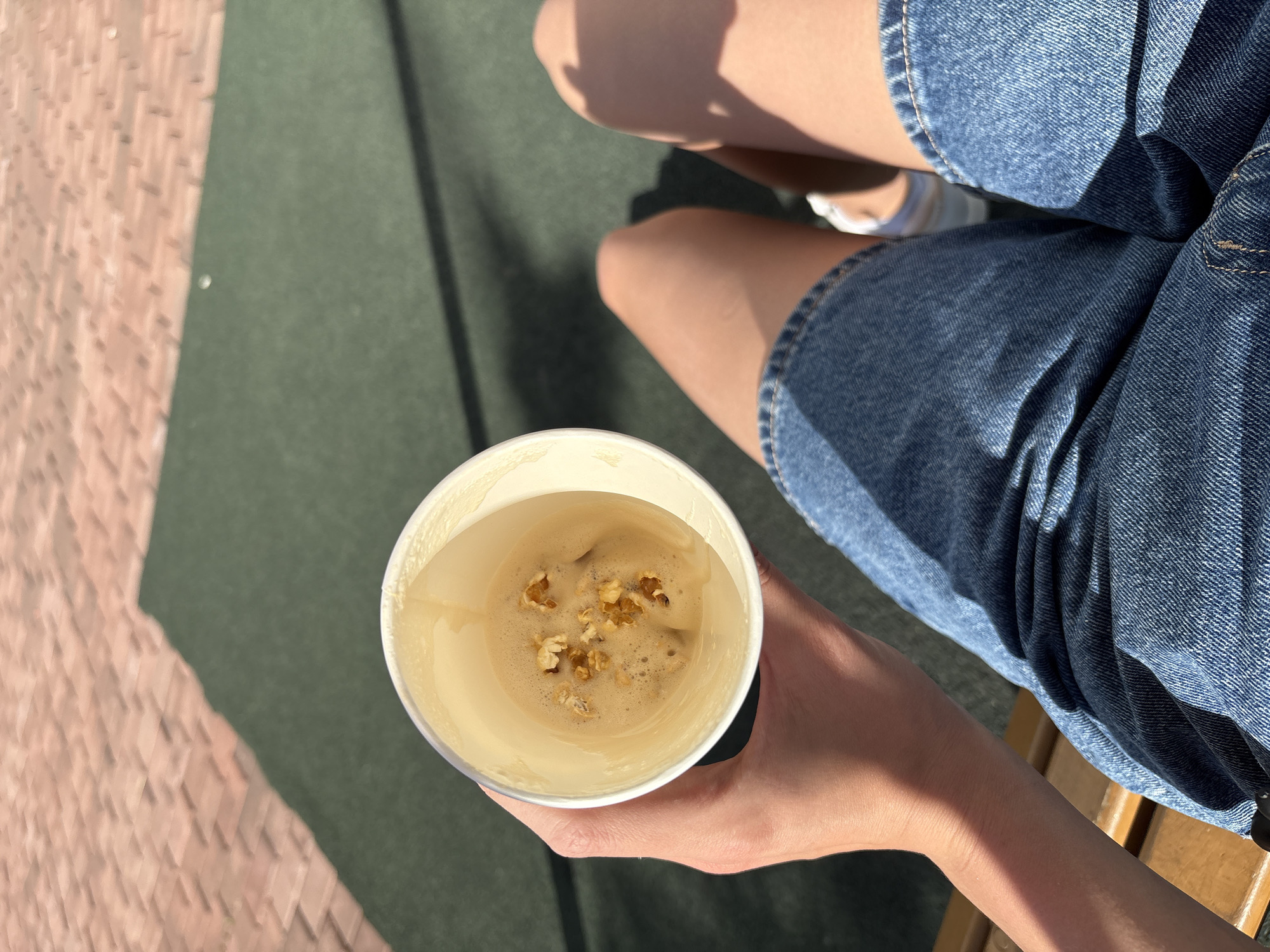 An overhead, close-up outdoor shot captures a person holding a white paper cup containing a light brown beverage. Small pieces of what appear to be popcorn or puffed grains float on the surface of the drink, which has a frothy top. The person's bare legs and denim shorts are visible in the background, seated on a wooden bench, partially visible on the right.
The ground beneath is split between two distinct surfaces: a reddish-brown, possibly tiled or brick, area on the left, and a dark green, uniform surface (potentially asphalt or rubber) on the right. Bright sunlight casts sharp shadows from the person's legs and the cup, indicating a sunny day, likely mid-morning to late afternoon. The scene conveys a casual, relaxed atmosphere, suggesting a moment of leisure outdoors, possibly in a park or public space in Krasnoyarsk, Russia. No discernible text is visible.