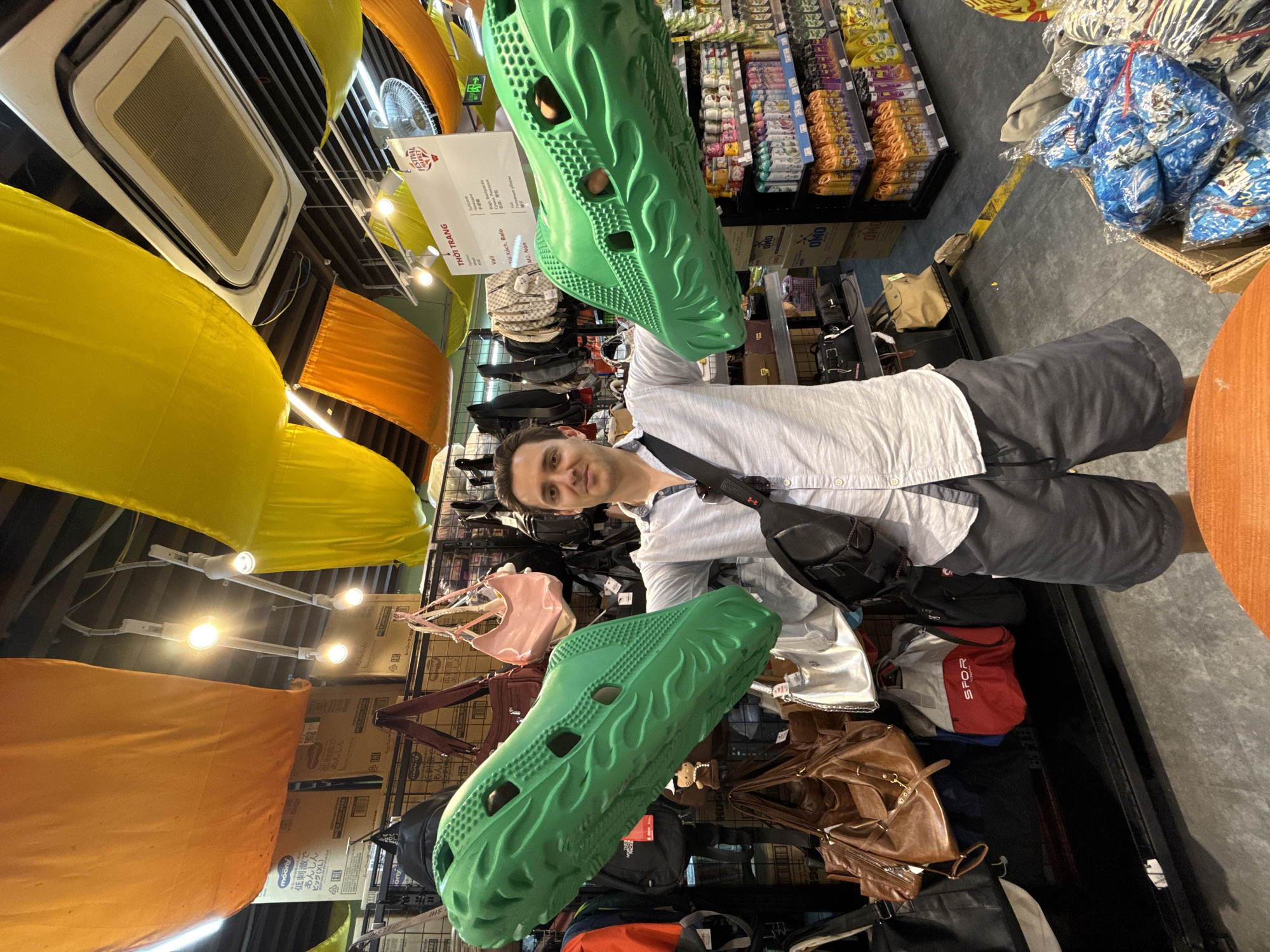 The image depicts a man standing in a retail environment in Bắc Nha Trang, Vietnam. He is holding a pair of oversized, green novelty sandals. The store appears to sell a variety of goods, including food products, purses, and luggage. The interior is decorated with hanging yellow and orange fabric. Signs in Vietnamese are visible.