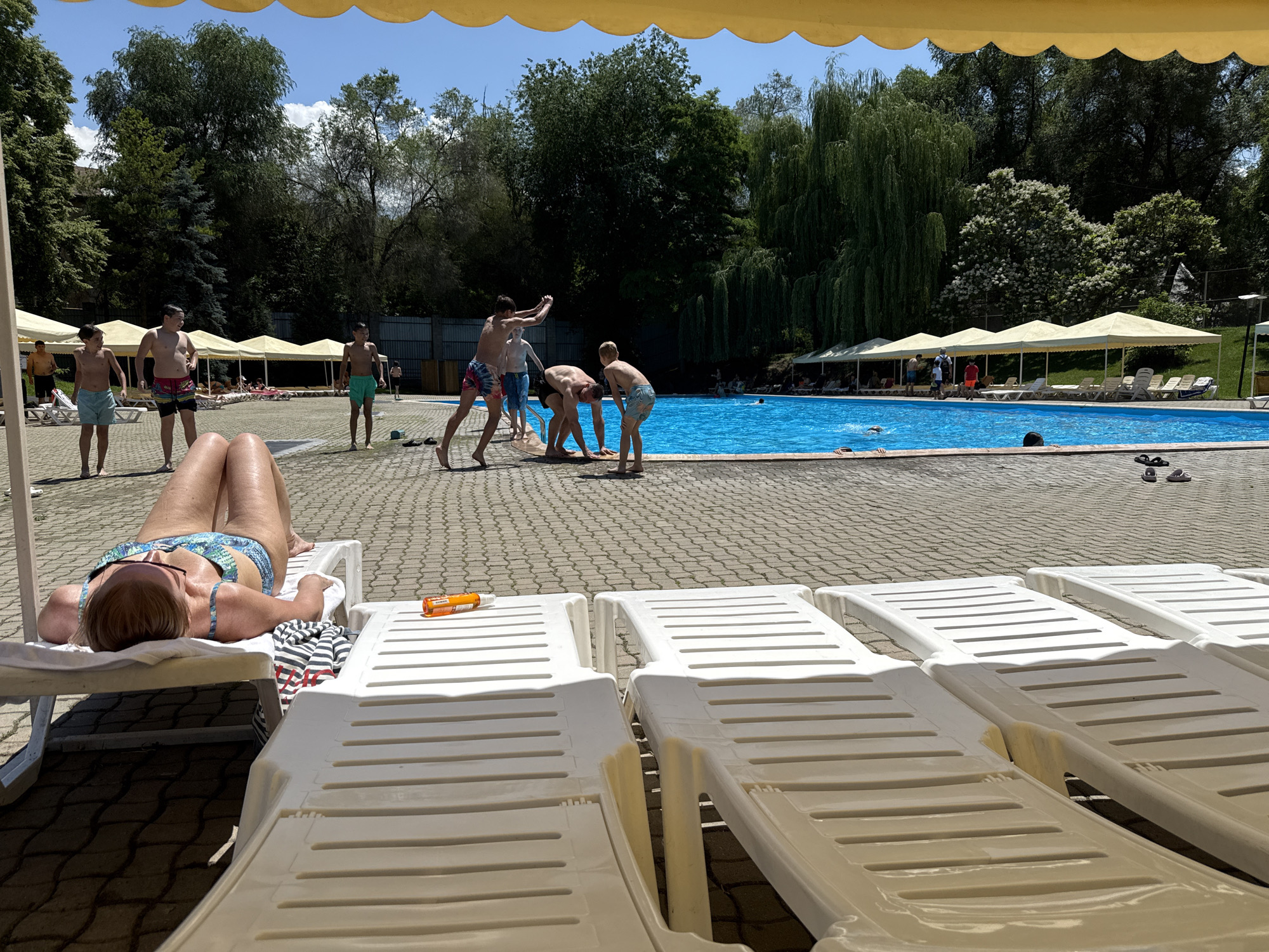 A sunny day at a swimming pool complex. The foreground features several empty white plastic lounge chairs, with one occupied by a woman in a blue and green bikini, lying on her back and sunbathing. Next to her is a towel with a striped pattern and a bottle of sunscreen. Beyond the lounge chairs, a paved area surrounds a large, bright blue swimming pool. Several people, mostly young males, are visible near the pool, some playing in the water and others standing on the edge. In the background, there are tents providing shade over more lounge chairs, lush green trees, and a fence. The environment suggests an outdoor leisure facility, likely a water park or resort, in Almaty, Kazakhstan.