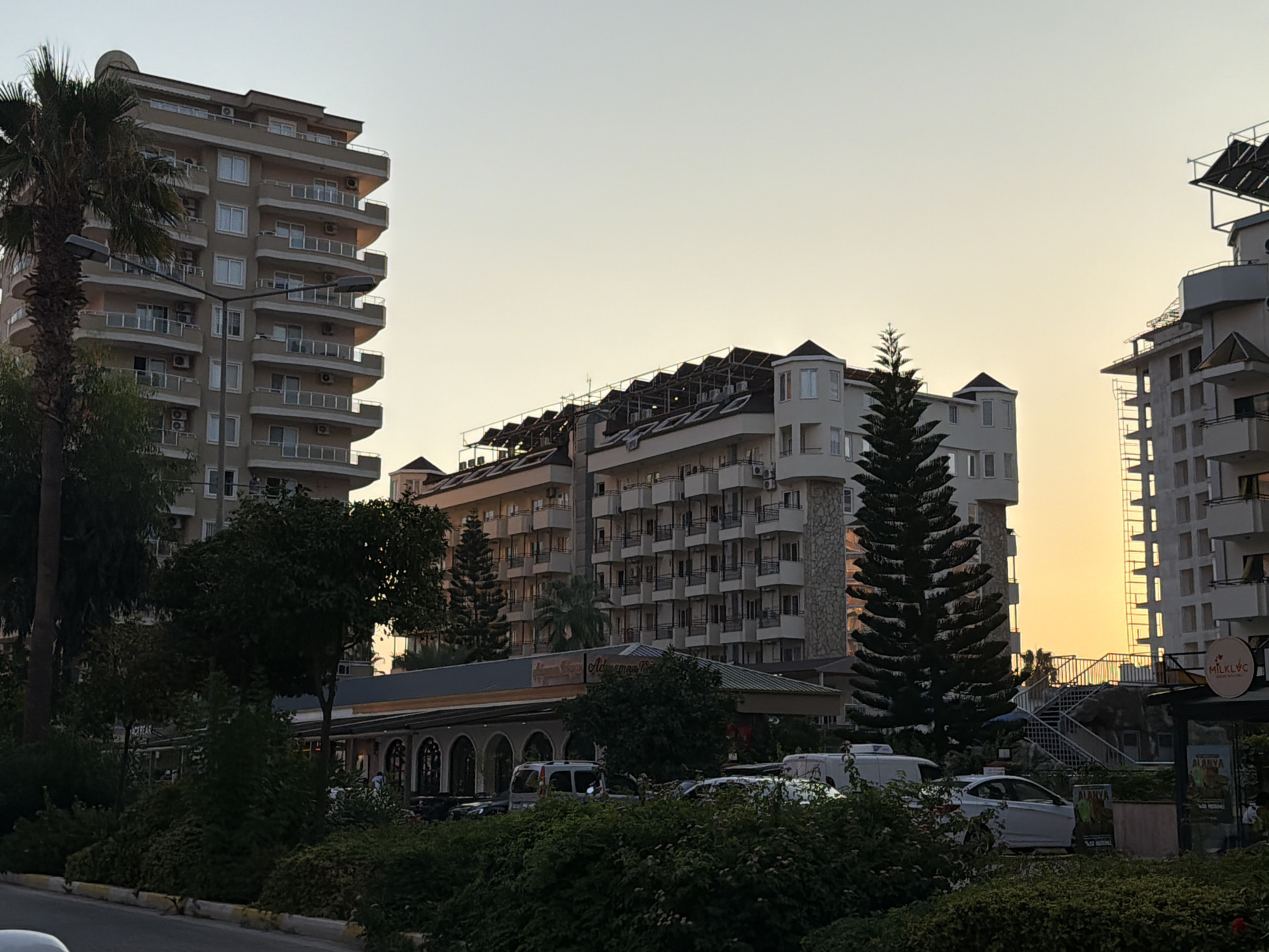 The image shows a street scene in Alanya, Turkey, with several buildings and lush greenery. In the foreground, there is a sidewalk and a street with parked cars, including a white sedan and a white van. A dense line of bushes and trees forms a natural barrier between the street and the buildings. Behind the greenery, a long, low-rise building with arched windows and a covered seating area is visible, possibly a restaurant or cafe. Above this, several taller buildings dominate the skyline. On the left, a tall, light-colored apartment building with numerous balconies and air conditioning units stands next to a palm tree and streetlights. To the right of the low-rise building, a darker, modern-style building with multiple balconies and windows is partially visible. Further to the right, another light-colored building with a distinctive turret-like structure on its corner and a prominent conical evergreen tree in front of it. In the background, a large, multi-story hotel or apartment complex features numerous balconies and solar panels on its roof. The sky is clear with a warm, golden hue, indicating either sunrise or sunset. The overall impression is a vibrant urban environment with a touch of tropical atmosphere.