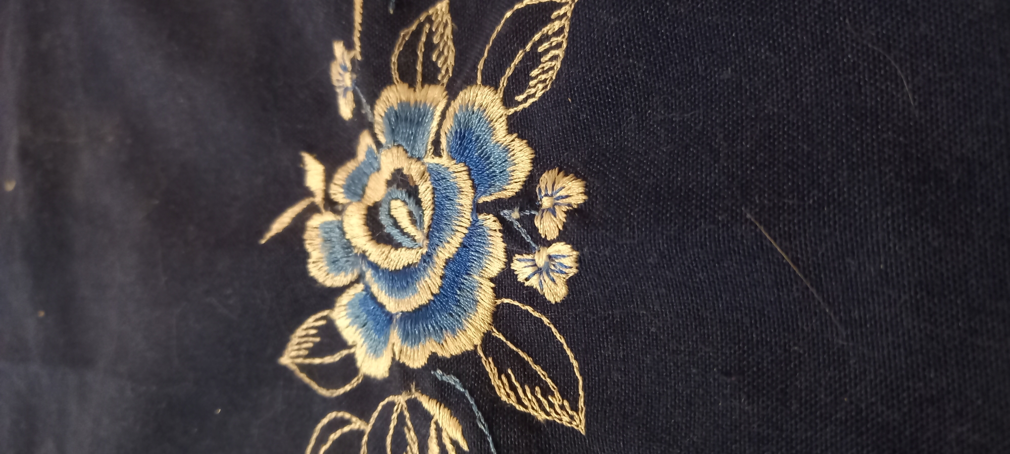 A close-up, high-angle shot shows a dark blue fabric with an embroidered blue and gold rose. The embroidery is detailed, with layered petals in shades of blue and outlined in gold thread. Smaller gold embroidered leaves and two small gold clusters of flowers are also visible. The fabric appears to be textured, possibly linen or cotton. There are a few stray strands of lint on the fabric. The location of this image in Kryvyi Rih, Ukraine, is unknown and cannot be determined from the visual information provided.