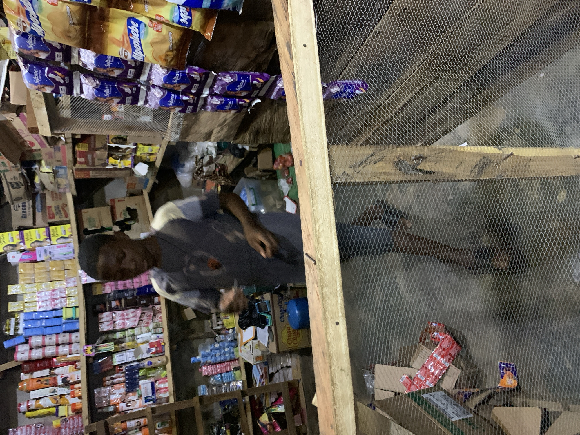 The image, rotated 90 degrees clockwise from its upright position, depicts the interior of a compact and densely packed shop or kiosk, likely situated in Kabba, Nigeria. The scene is rich with various consumer goods, neatly yet tightly arranged on wooden shelves and displays.
In the left-center of the frame, a male individual is visible, wearing a grey short-sleeved top. His head is slightly turned away from the camera, and his right hand is engaged with items on a lower shelf, suggesting he is working or attending to the store.
The shop is abundantly stocked with a wide array of packaged products. Prominently displayed at the top are multiple rows of purple-packaged "Softcare" items, alongside golden "Alpenliebe" branded products with the word "Creamy" also visible. Other discernible text on packaging includes "Chupa Chups" on boxes, "GLUCOS," "Vita," and a partial "Maga." These items range from confectioneries and snacks to what appear to be personal care products.
The structure of the shop features robust wooden framing for the shelves and a notable wire mesh partition that covers a significant portion of the right side of the image (when correctly oriented). This mesh likely functions as a security measure or to separate different areas of the store. The lighting within the shop is artificial, illuminating the merchandise. The overall impression is that of a bustling local commerce environment, characteristic of everyday life in Kabba, Nigeria.