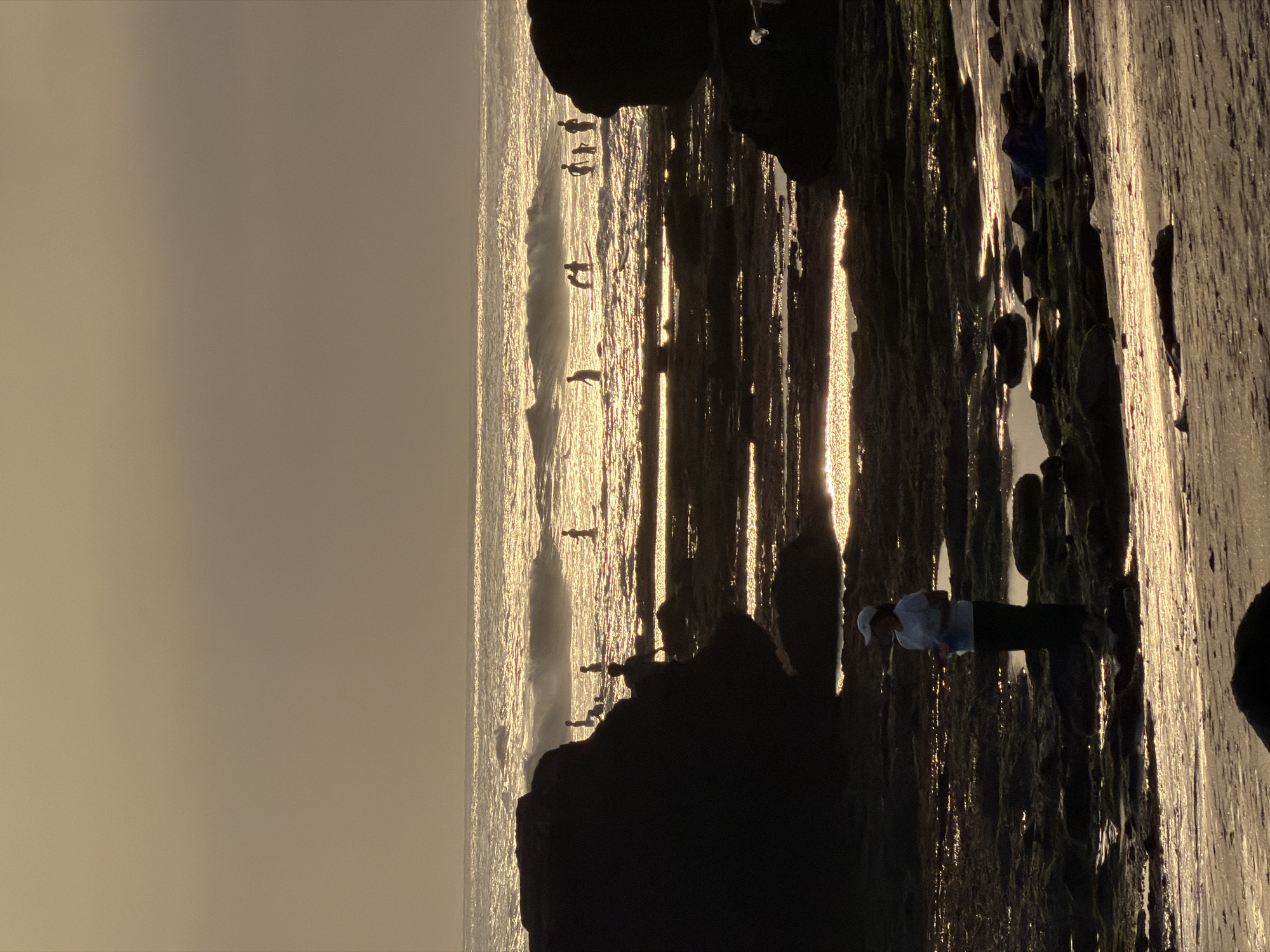 A photograph taken at dusk in North Kuta, Indonesia, captures the receding tide on a rocky beach. In the foreground, a person stands on the wet sand, silhouetted against the shimmering ocean. Beyond them, the water is dotted with numerous individuals, some wading, others standing or walking, all appearing as dark figures against the golden glow of the setting sun. Small waves break in the distance, reflecting the warm light. The rocky shoreline in the mid-ground features pools of water left by the tide, also reflecting the sunset. The overall scene is bathed in a soft, diffused light, creating a tranquil and atmospheric view of a popular coastal area.