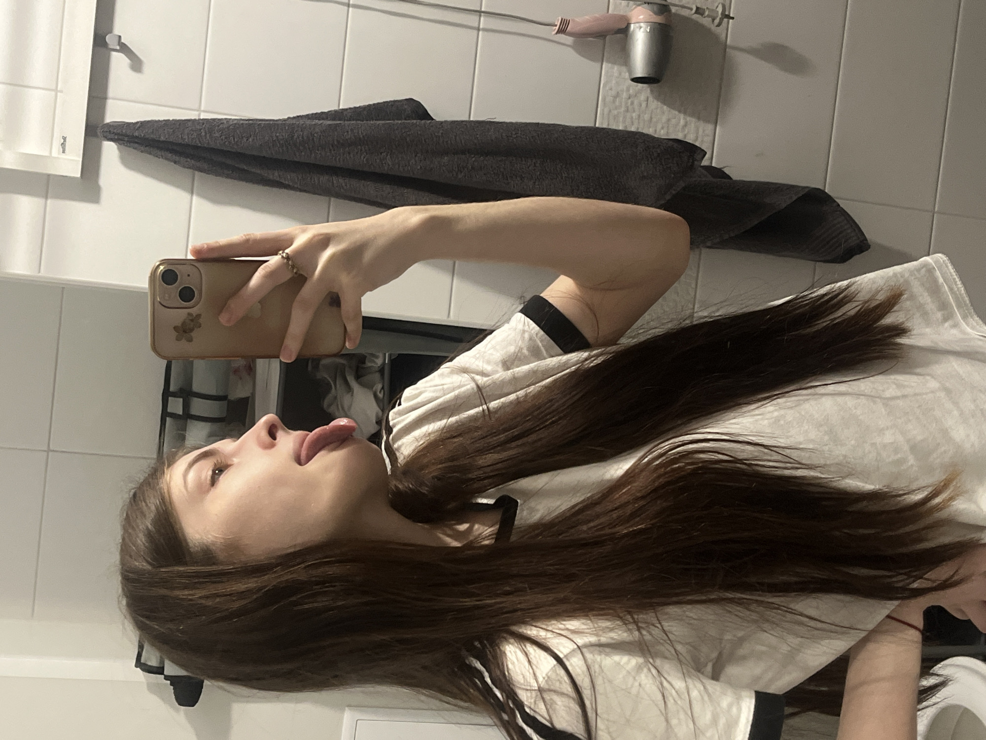 A young female with long, dark brown hair is captured in a mirror selfie, seen in profile, with her head tilted slightly back and to her left, tongue extended. She wears a white t-shirt with black trim on the collar and sleeves. Her right hand, adorned with a beaded ring on the ring finger, holds a gold-colored smartphone featuring a floral design near its dual camera lenses. Her left wrist, bearing a red string or bracelet, is partially visible in the lower-left corner. The setting is an indoor bathroom, characterized by white tiled walls with horizontal grout lines. Visible in the background are a dark grey textured towel hanging on a silver wall hook, a rectangular mirror reflecting the ceiling, and a pink and silver hairdryer mounted below a hook on the right. An open shelf or cabinet containing folded items is partially visible behind the subject's head. A portion of a toilet rim is faintly visible at the bottom left. The specific location in the city of Saint Petersburg, Russia, cannot be determined from the visual information presented.