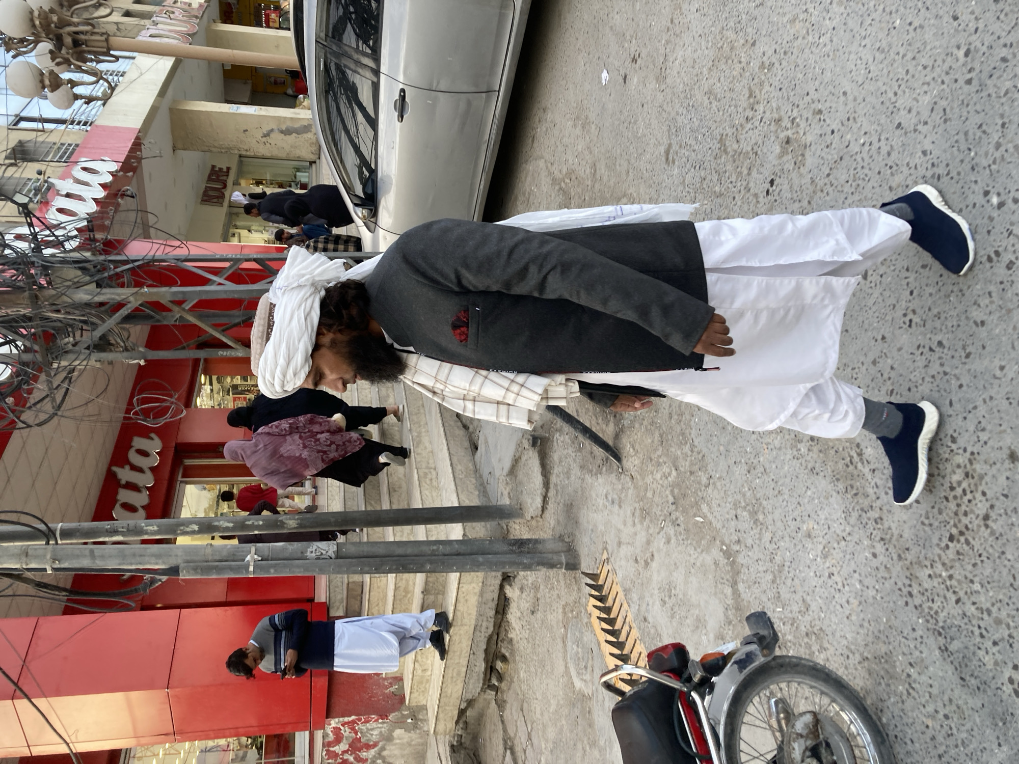 The image shows a scene in Quetta City Tehsil, Pakistan. A man wearing traditional attire, including a white turban and shalwar kameez, with a dark gray vest, is standing on a sidewalk. Behind him, a silver car is parked near a building with a red facade and the word "Bata" in large white letters. Several other people are visible on the steps leading into the building. To the man's right, a motorcycle is partially visible. Overhead, a complex network of utility wires is present. The setting appears to be an urban commercial area.