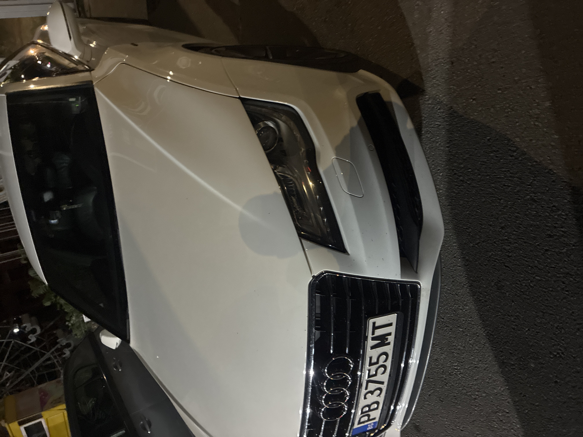 A white Audi car is prominently featured, parked outdoors at night in what appears to be Asenovgrad, Bulgaria. The image, captured from a slightly elevated angle, primarily shows the front and left side of the vehicle, which is positioned with its front facing the right side of the frame. The car's sleek, white body reflects artificial light sources, indicating a nighttime setting.
The iconic four-ring Audi logo is clearly visible on the chrome grille. Directly adjacent to the logo, the Bulgarian license plate "PB 3755 MT" is displayed, with the "BG" country code also visible, confirming its registration in Plovdiv Province. The car's left headlight is visible and appears to be illuminated or reflecting light. Parts of the driver's side door, windshield, and a side-view mirror are also discernible.
The car is situated on an asphalt surface, visible in the foreground and extending to the right. The background is dark and out of focus, showing indistinct shapes of what might be buildings or other structures, and a partial view of another dark-colored vehicle to the lower left. No people are present in the image, and the car appears to be stationary. The scene captures a quiet, late-night moment with the parked vehicle.