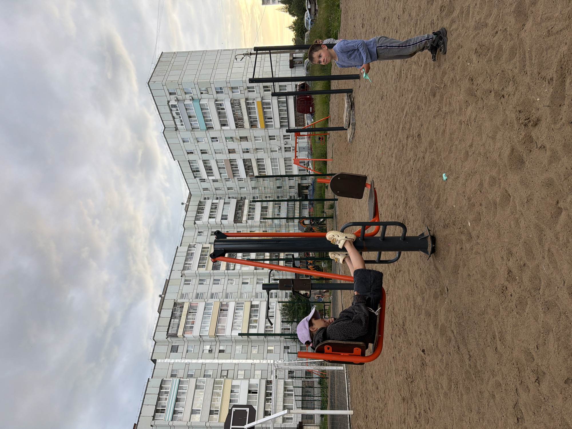 The image depicts an outdoor playground with two children. The setting is a sandy area in front of a large, multi-story apartment building, characteristic of Chaikovsky, Russia. The building is a light gray color with numerous windows and balconies. The sky is overcast with soft clouds, suggesting it might be late afternoon or evening.
In the foreground, a child wearing a purple cap, a dark patterned hoodie, and black shorts is seated on an orange exercise machine. Their feet are resting on a bar, and they appear to be relaxed or engaged in using the equipment. To the right and further back, another child is standing near a parallel bar structure. This child is wearing a blue long-sleeved shirt and gray pants with stripes, and they are holding a small light blue object. Their posture suggests they are either resting or have just finished an activity.
The playground equipment includes the exercise machine and the parallel bars, both appearing to be made of metal with orange accents on the exercise machine. The sand surface of the playground is visible throughout the image. The overall atmosphere seems calm and everyday, with the children enjoying their time outdoors. There is no visible text in the image.