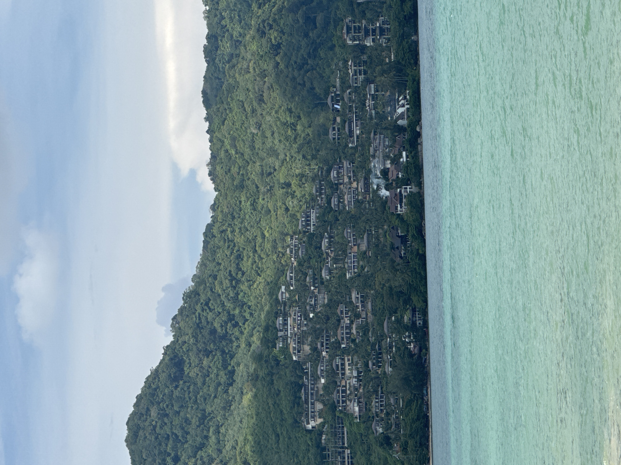 The image depicts a scenic view of a lush green mountain slope dotted with numerous buildings, cascading down towards a tranquil, turquoise sea. The buildings appear to be modern villas or resort accommodations, integrated into the natural landscape. The dense foliage of the tropical mountain covers much of the structures, creating a harmonious blend of architecture and nature.
The sea exhibits gentle ripples, suggesting calm weather conditions. The water's clarity allows for a glimpse of the seabed in some areas, further enhancing the idyllic tropical setting. The sky above is partly cloudy, with patches of blue sky interspersed with white clouds. A bright, luminous band of light, possibly a sunbeam or a reflection, cuts vertically through the sky on the left side of the image.
The scene is likely set in the late afternoon or early evening, given the soft lighting. The location appears to be a popular tourist destination, possibly Ban Bang Thao in Thailand, known for its beautiful beaches and resorts nestled amidst hills. There is no visible text in the image. The overall mood conveyed is one of serenity, luxury, and natural beauty.