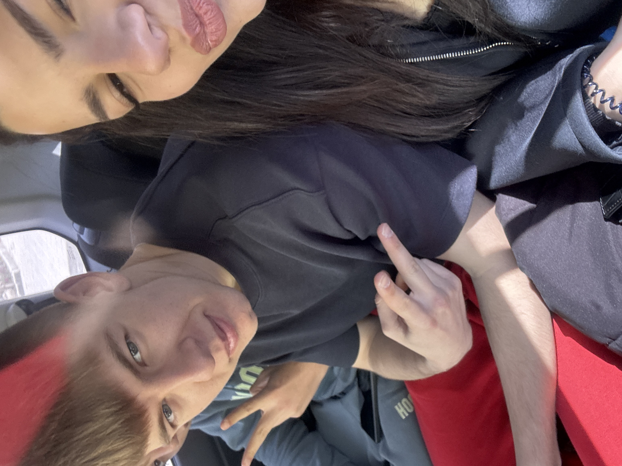 A selfie captures two individuals in what appears to be a vehicle's interior. The image is oriented at an angle, with the top of a woman's head visible at the top of the frame. She has dark hair, fair skin, and pink lipstick, and is wearing a black jacket with a visible zipper. Below her, a male subject is positioned more centrally in the frame. He has light hair with a red streak, fair skin, and is wearing a black t-shirt. He is displaying the middle finger gesture with his right hand. A peace sign is visible in the bottom left corner, and part of a red garment can be seen. The location is unspecified.