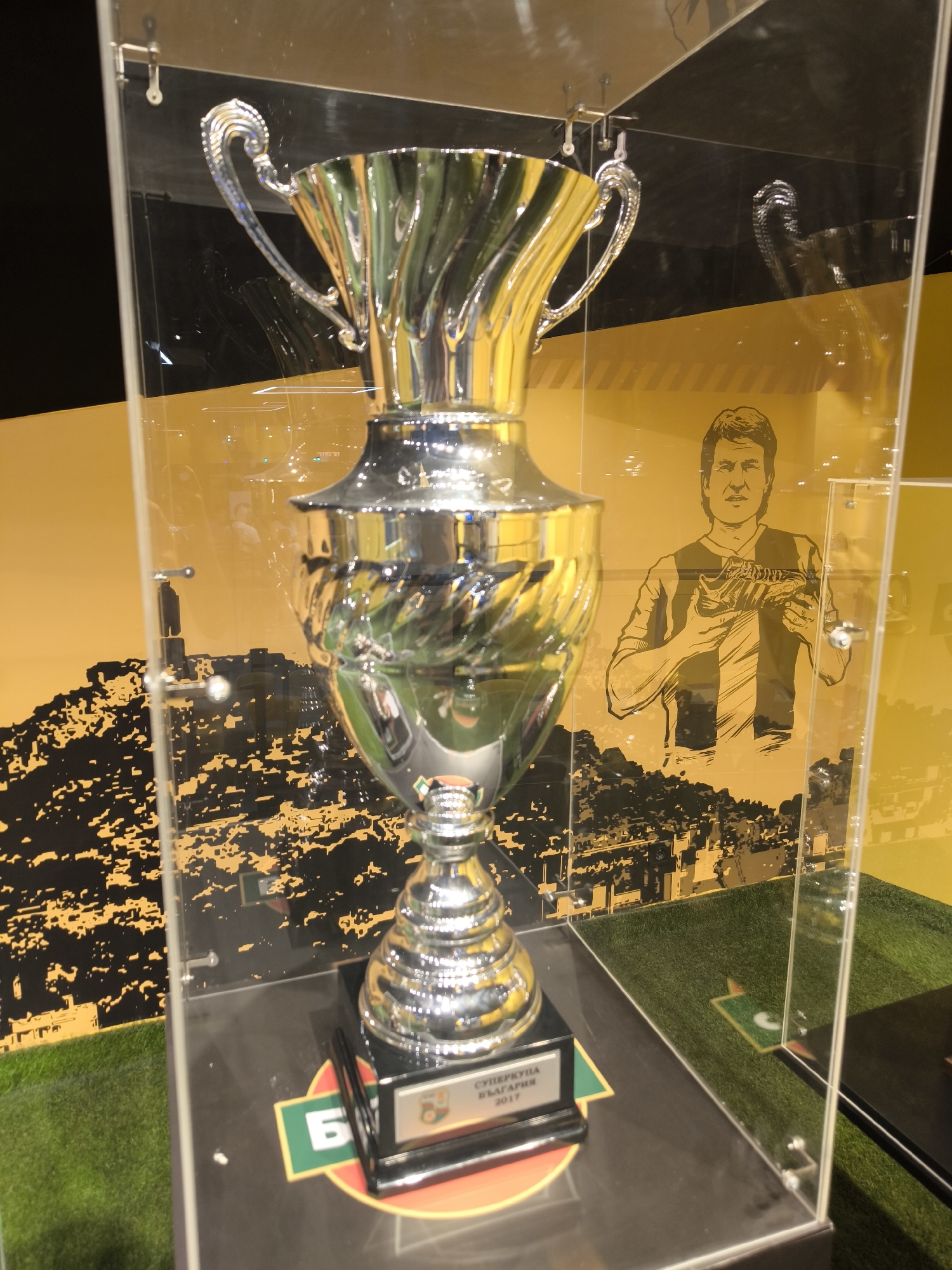 The image shows a silver trophy displayed in a glass case. The trophy has intricate detailing and is mounted on a black base with a plaque that reads "СУПЕРКУПА БЪЛГАРИЯ 2017". Behind the trophy, there is a yellow wall with a graphic of a cityscape and a drawing of a man holding a football. The display case appears to be situated within a museum or exhibition. The artificial grass floor and the overall presentation suggest a sports-related theme, possibly related to Bulgarian football. The scene is indoors, under artificial lighting. No people are visible in the immediate vicinity of the trophy. The location in the city of Plovdiv, Bulgaria, is not directly evident from the image itself, but based on the trophy's inscription, it likely commemorates a football championship in Bulgaria, and Plovdiv is a major city in Bulgaria that hosts sporting events.