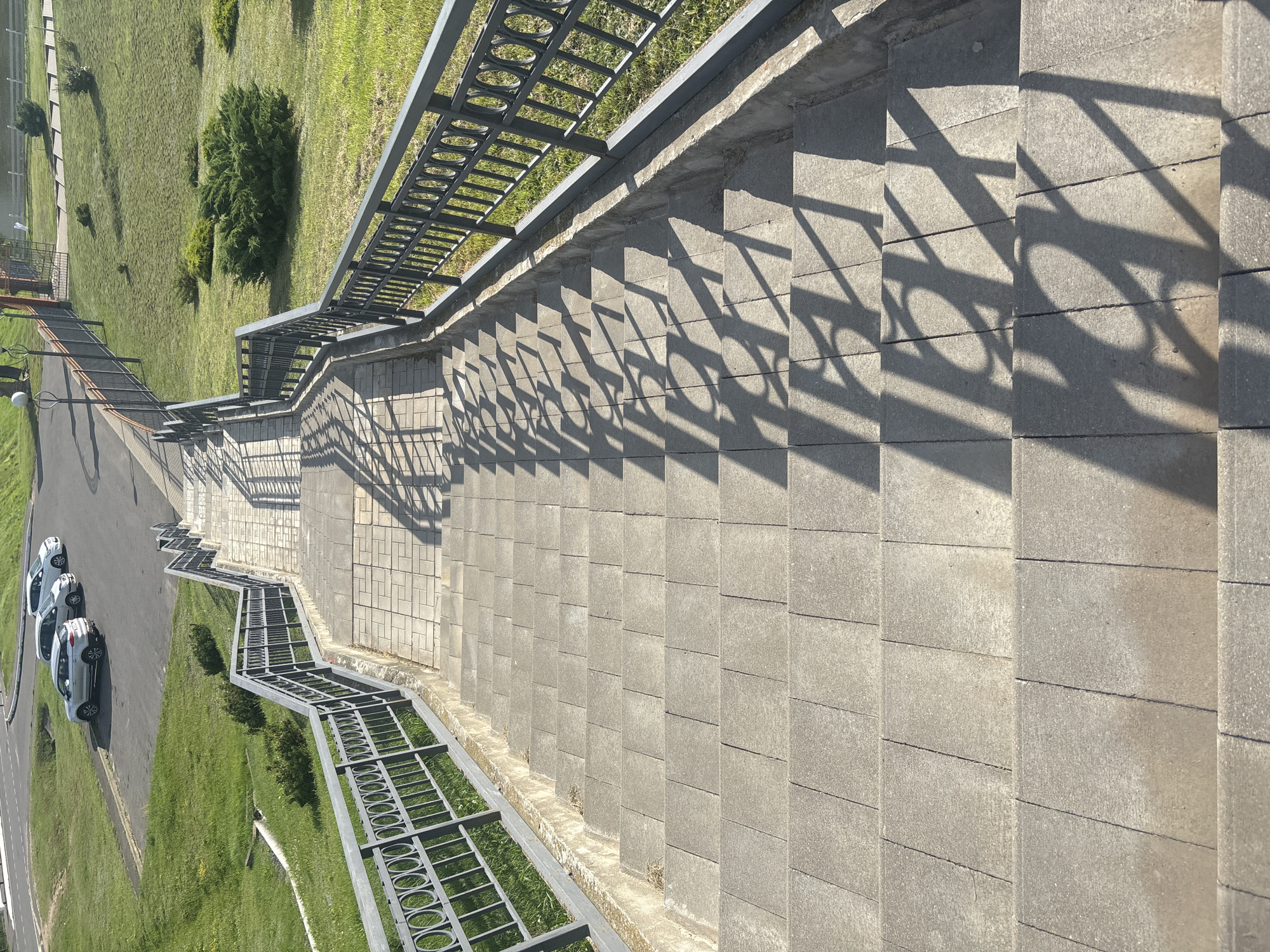 The image shows a downward view of a wide, gray concrete staircase with metal railings on both sides. The staircase is flanked by a grassy slope on the left and a manicured lawn with shrubs on the right. In the distance, at the top of the stairs, there is a paved area with several cars parked, and a road. The sun casts distinct shadows of the railings onto the steps. The location is identified as Kapyĺ, Belarus.