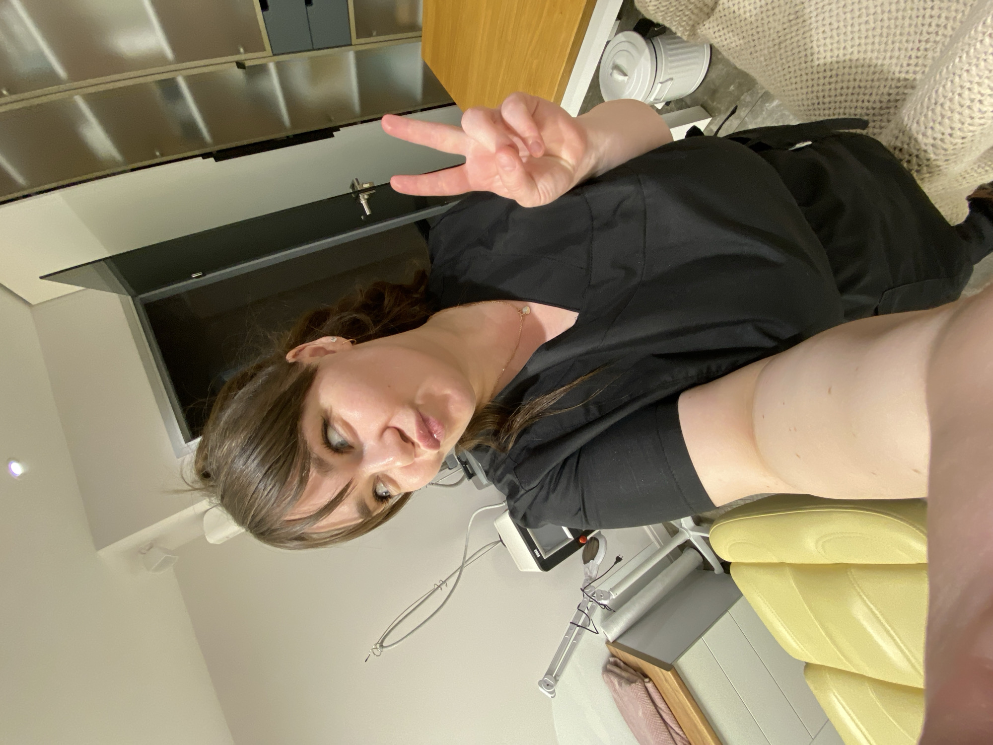 A woman is seated in a yellow medical chair, making a peace sign with her right hand. She has brown hair, and is wearing a black scrub top and a necklace. The background includes medical equipment, cabinets, a desk, and a waste bin. It is not possible to determine the exact location in Votkinsk, Russia based on the image.