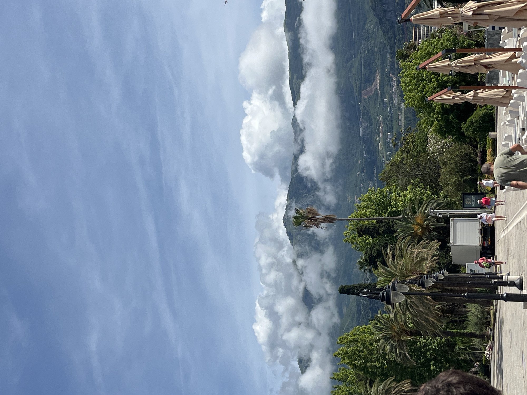 The image captures a scenic daytime view of a bustling promenade against a dramatic mountainous backdrop in Budva, Montenegro. The sky, predominantly clear light blue with wispy white clouds, occupies the upper left portion of the frame. Fluffy white clouds also partially obscure the lush green mountains that stretch across the middle ground, some clinging to the peaks and slopes, while a small bird is visible flying in the upper sky.
In the mid-right, a paved promenade or street is lined with several dark, ornate street lamps and numerous palm trees with varying frond textures. On the far right, a building features multiple closed, light-beige patio umbrellas or awnings, suggesting a restaurant or cafe. Below these umbrellas, white structures, possibly benches or parts of a terrace, are visible.
Several people are present on the promenade. In the lower right, the top of a person's head and hair are visible, suggesting they are in the immediate foreground. Further along the promenade, people are walking; one individual in a green shirt is seen from behind, and others in lighter clothing are scattered. A white truck or van is parked near the promenade, and next to it, a dark sign clearly displays the text "ZAYTOUN". The overall scene depicts a vibrant, sunny day in a coastal town, with the natural beauty of mountains and the activity of people enjoying the area.