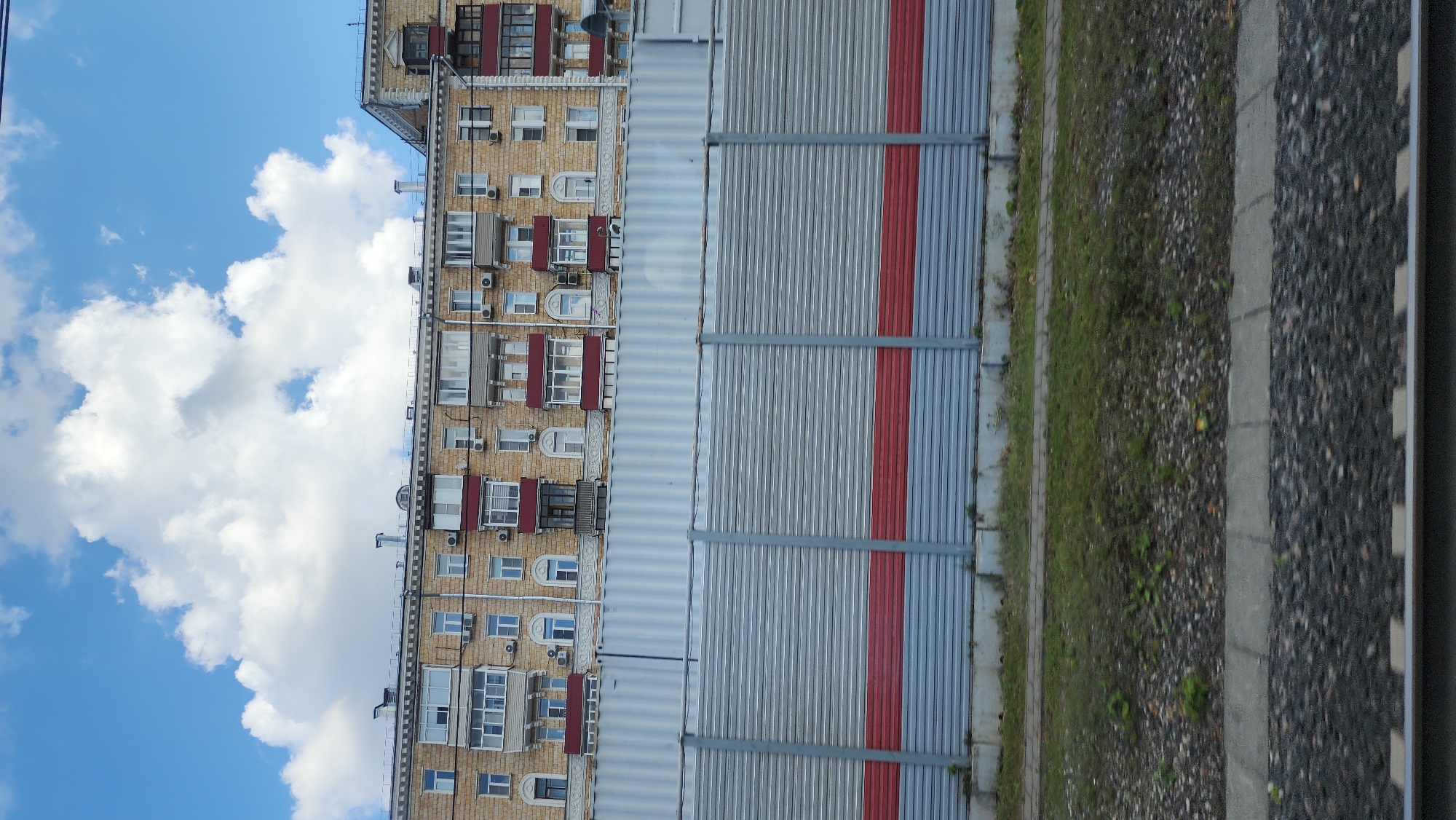The image shows a multi-story brick apartment building with numerous windows and balconies, under a cloudy blue sky. In front of the building is a tall, corrugated metal fence with red and gray bands. Below the fence is a grassy area with some gravel and a concrete path. In the foreground, a section of railroad tracks is visible. The scene appears to be located in the city of Khimki, Russia.