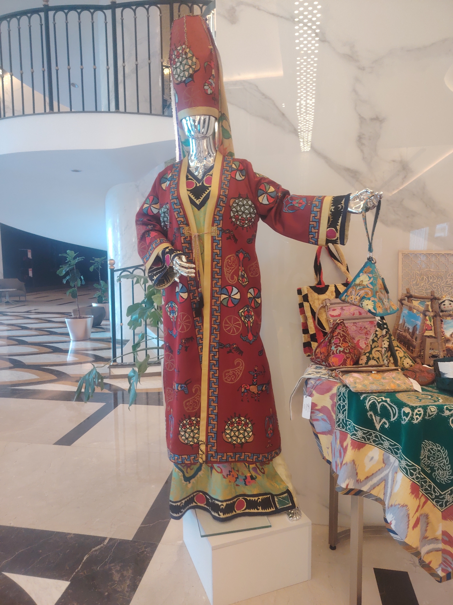 In Konigil, Uzbekistan, a mannequin is displayed wearing a traditional, richly decorated red robe with a tall, conical hat. The robe features intricate patterns in yellow, blue, and green, including circular motifs and depictions of horsemen. The mannequin's hands are silver. To the right of the mannequin, a table is covered with colorful textiles. Several patterned bags, including a large tote bag and smaller pouches, are arranged on the table, alongside what appear to be framed artworks and decorative dolls. The background shows a modern interior with marble flooring and a decorative ceiling light, and a metal railing. Several potted plants are visible in the left background, and the scene is illuminated by interior lighting.