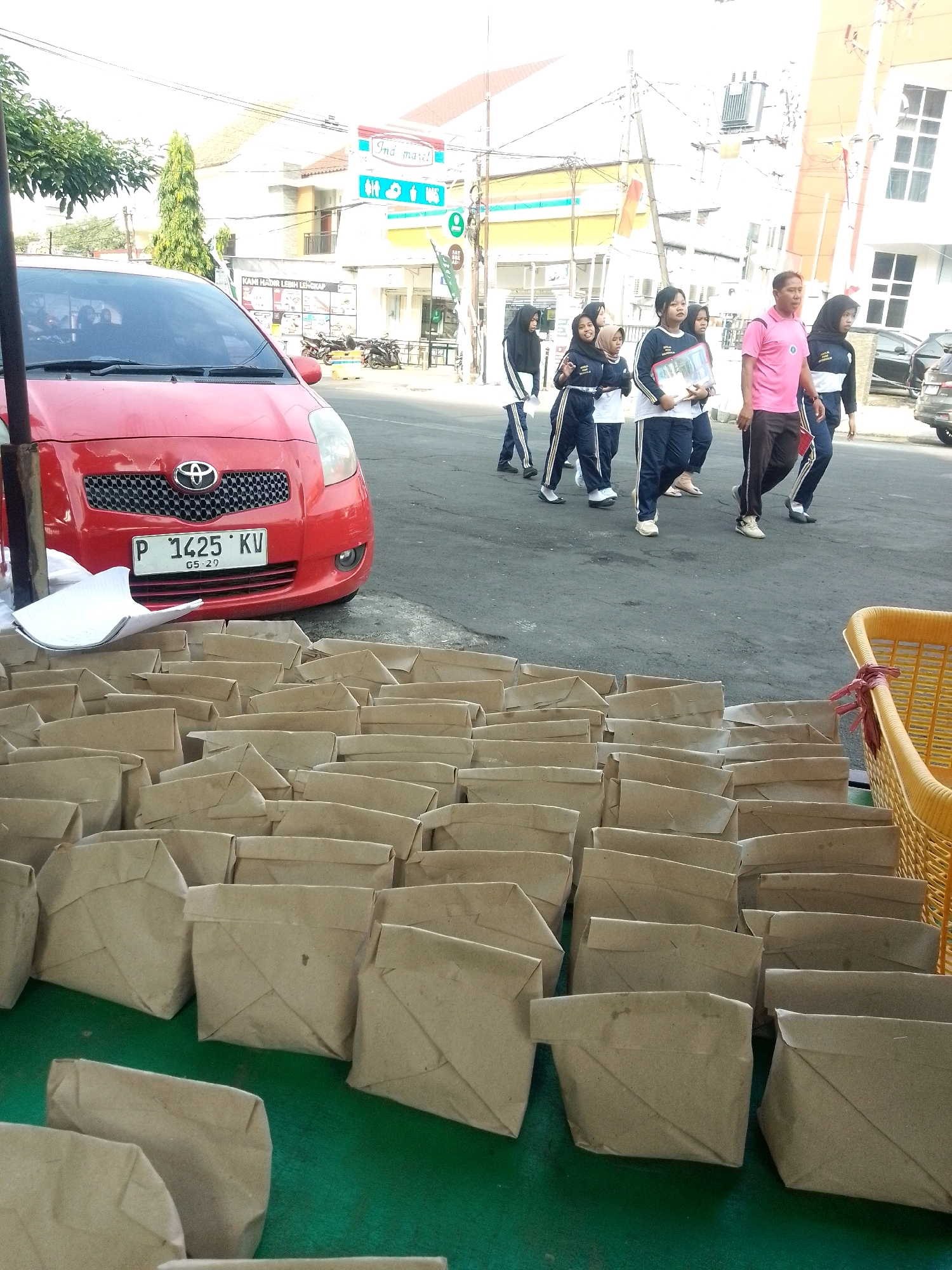 The image depicts an outdoor scene on a street in Jember, Indonesia, likely in front of commercial establishments. In the foreground, a large number of brown paper-wrapped packages are neatly arranged on a green surface, suggestive of a food distribution or sale. To the left, the front of a red Toyota car is visible, with a license plate reading "P 1425 KV 05-29."
In the midground, a group of people are walking on the asphalt road. The group consists of several young individuals, appearing to be students in school uniforms, and at least one adult male wearing a pink shirt and dark pants. The students are dressed in dark blue and white uniforms with some wearing hijabs. They are carrying folders and bags. Behind them, more people are walking.
The background shows buildings with signage, including one that reads "Indomaret," a common convenience store chain in Indonesia. There are also utility poles, wires, and trees, indicating an urban environment. The overall lighting suggests it is daytime. The scene appears to be a busy street, potentially near a school or a public gathering area.